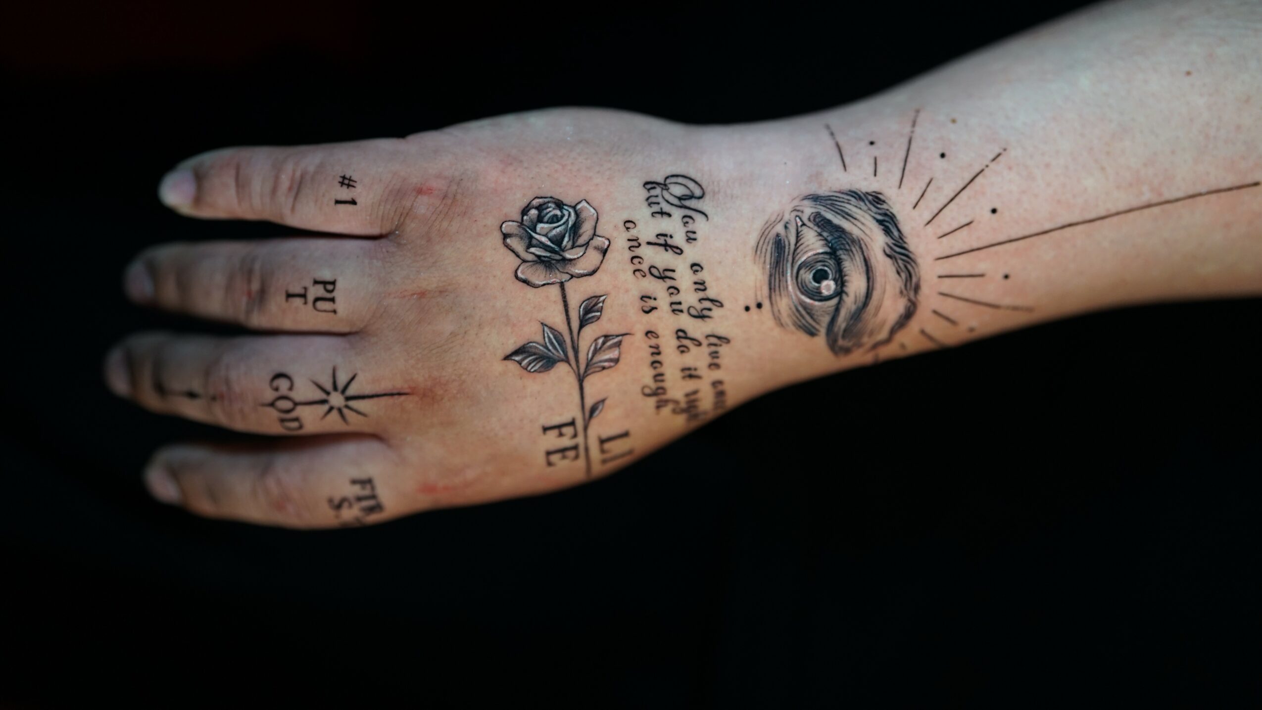 Tattooed hand with an eye, rose, and text, against a black background.