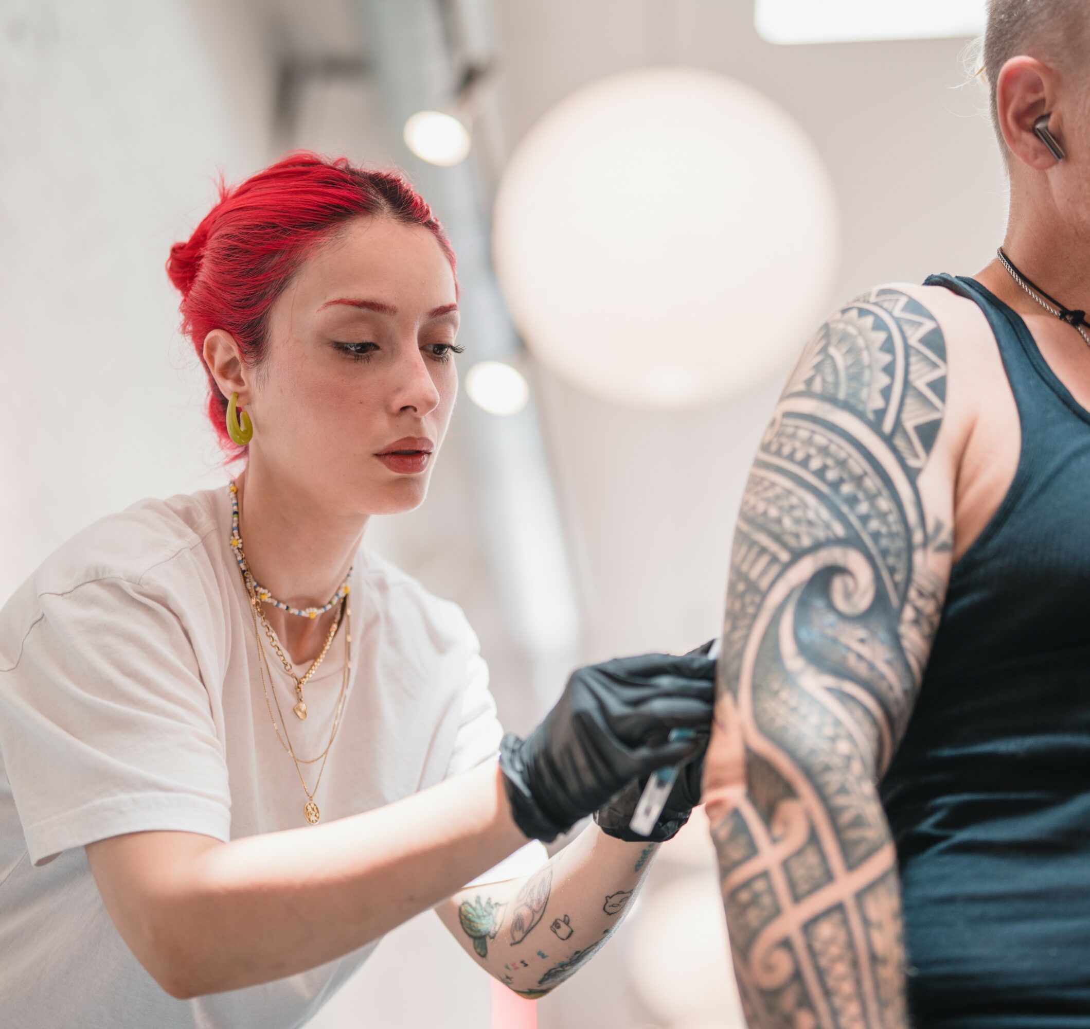 Tattoo artist with red hair and black gloves works on a person's arm tattoo in a studio.