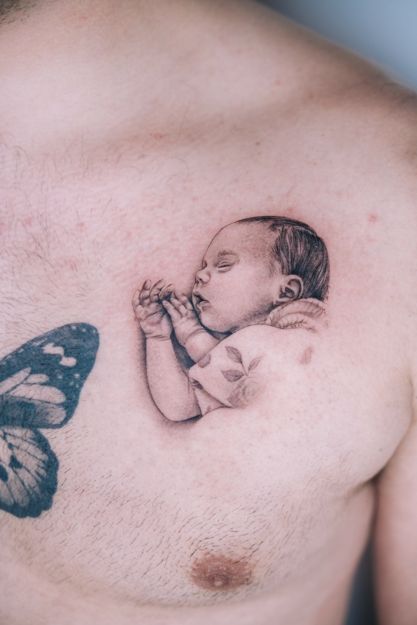 Tattoo of a sleeping baby on a person's chest, with a butterfly tattoo partially visible on the left.