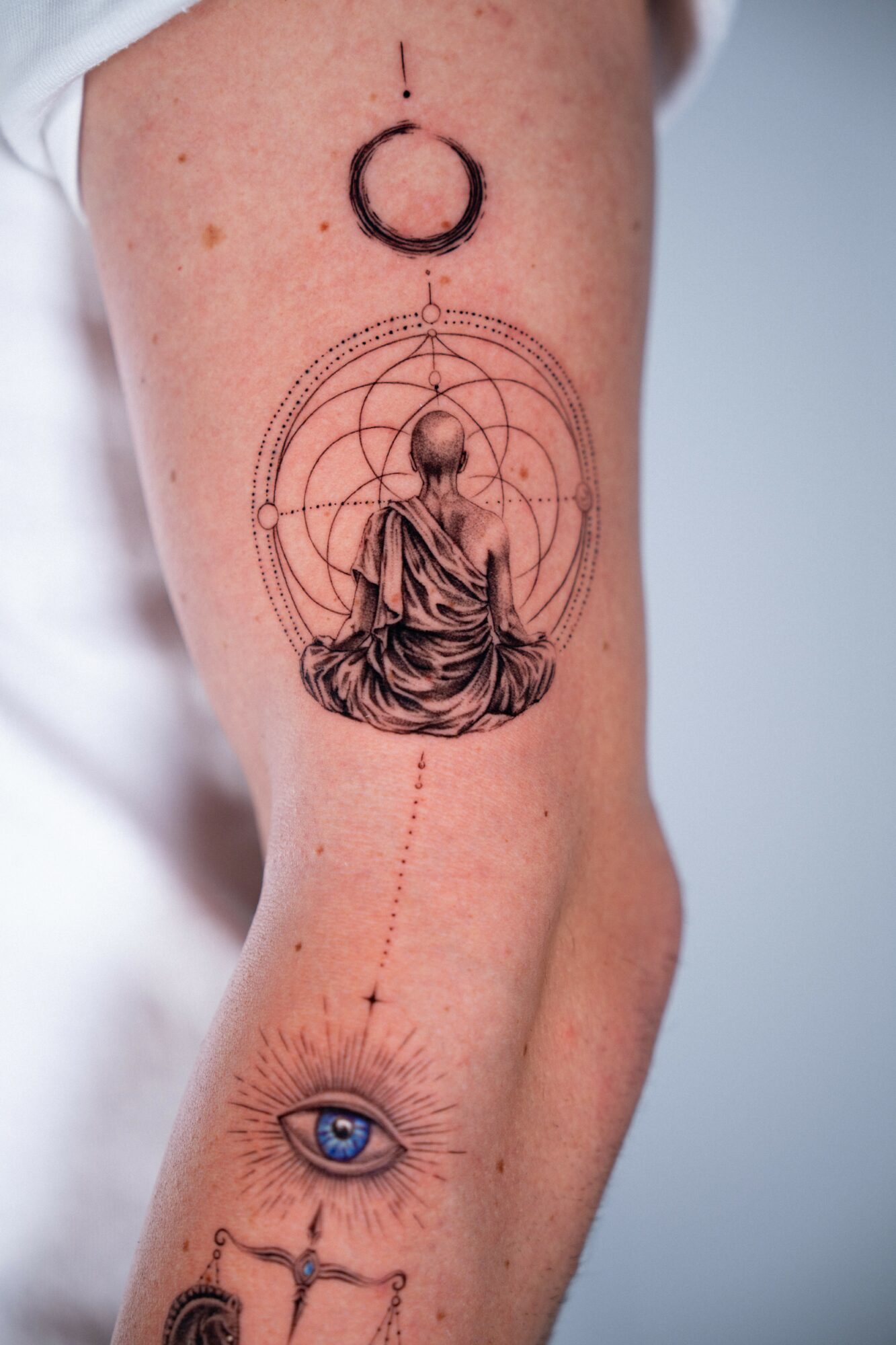 Tattoo of a meditating figure with geometric patterns, a moon, and an eye on a person's arm.
