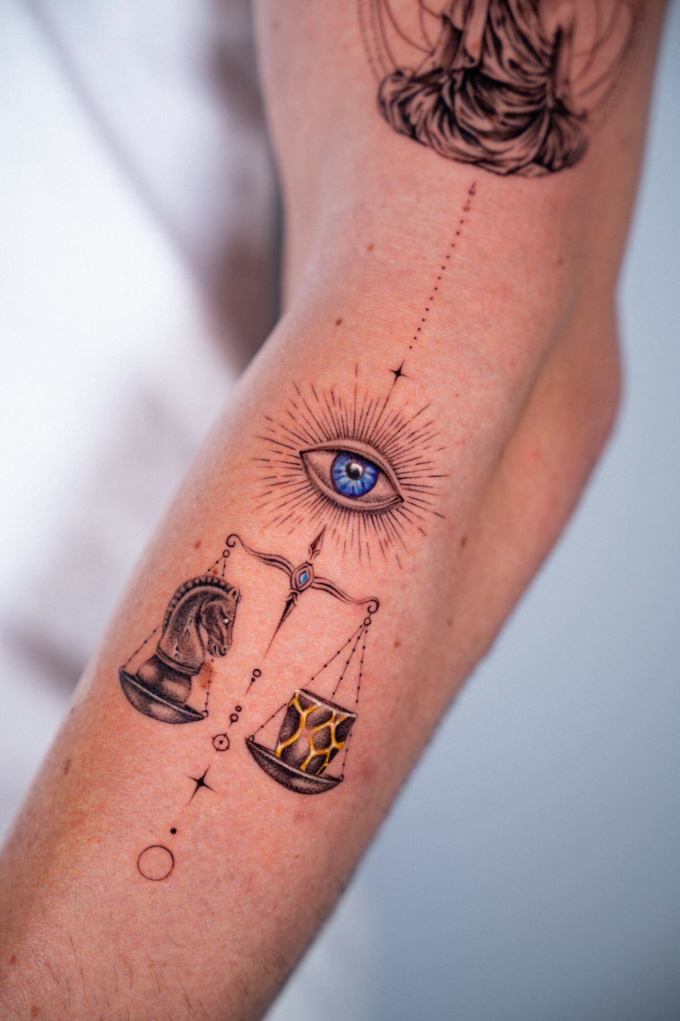 Tattoo of an eye with rays, a balance scale, and a lantern on a person's arm.