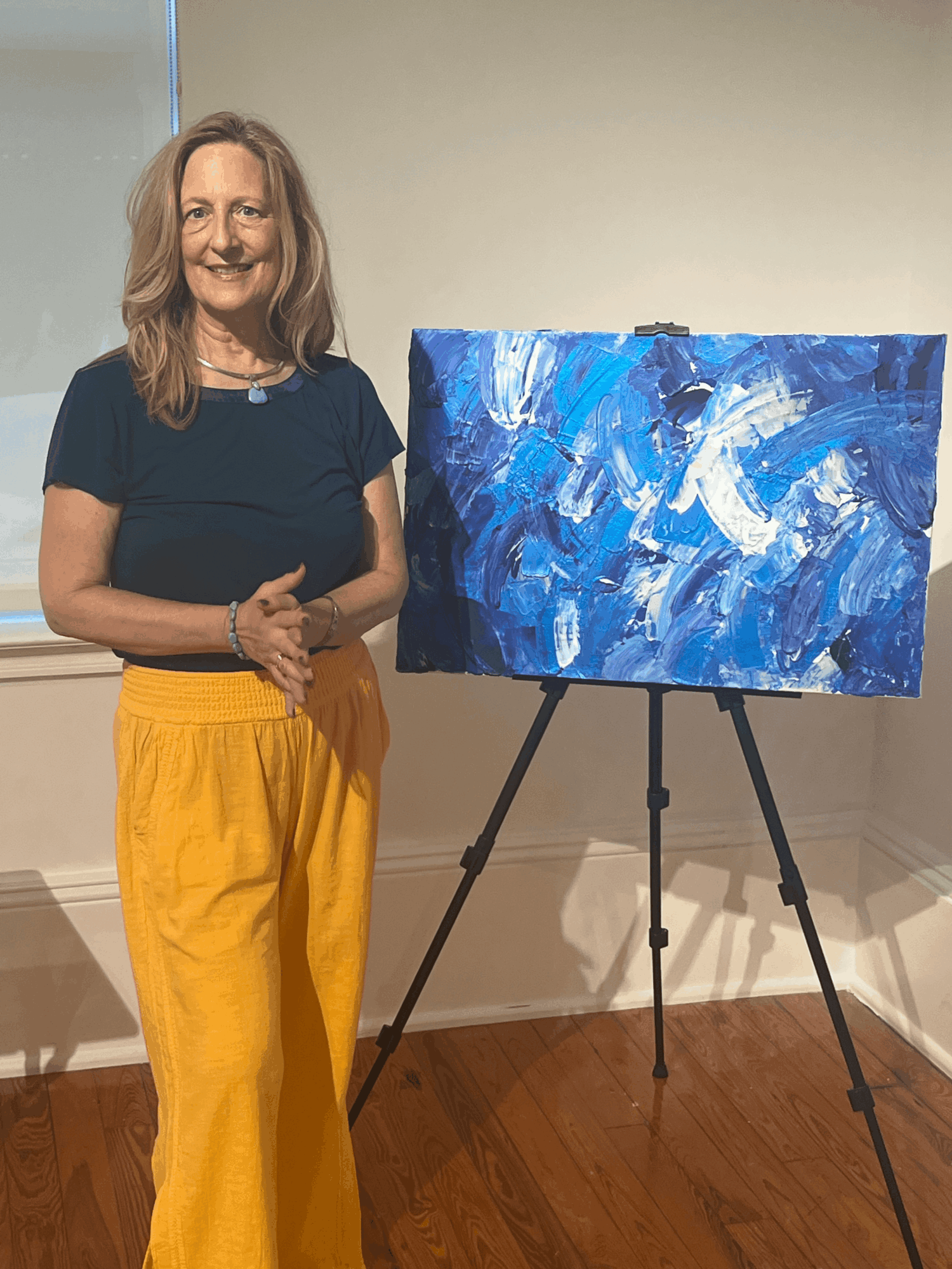 Woman with shoulder-length hair in a black shirt and yellow pants standing next to an abstract blue painting on an easel.