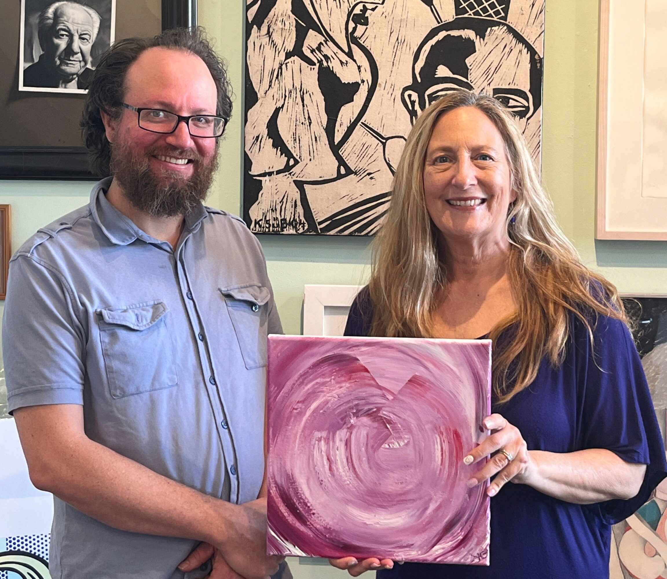 Two people, a man with glasses and a woman with long hair, hold a pink and purple abstract painting, smiling.
