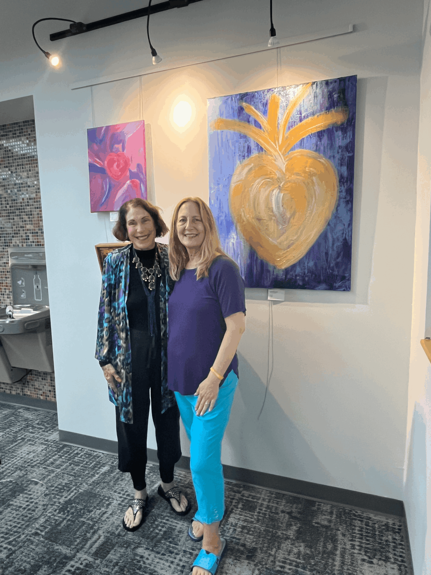 Two women stand in front of colorful abstract paintings on a gallery wall, smiling at the camera.