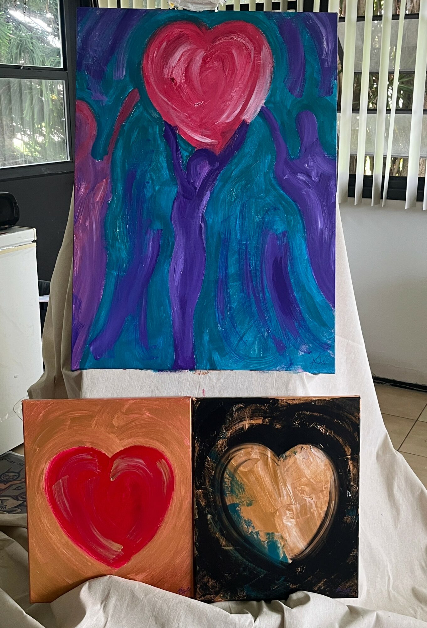 Three paintings with hearts, one large and two smaller, displayed on a stand indoors, with window blinds in the background.