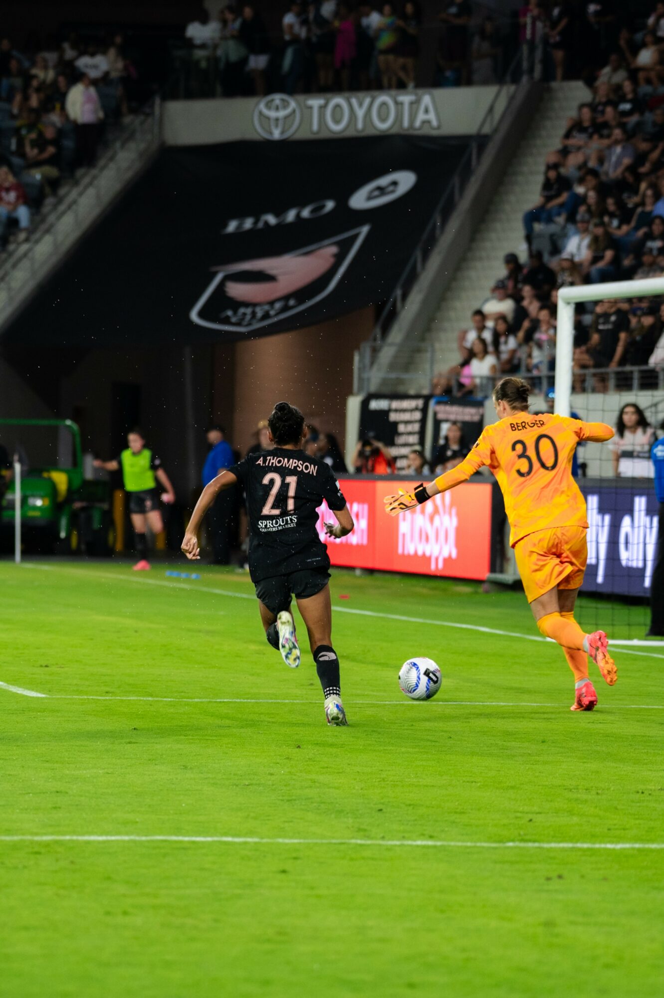 Two soccer players chase the ball on a green field, with spectators and advertisements in the background.