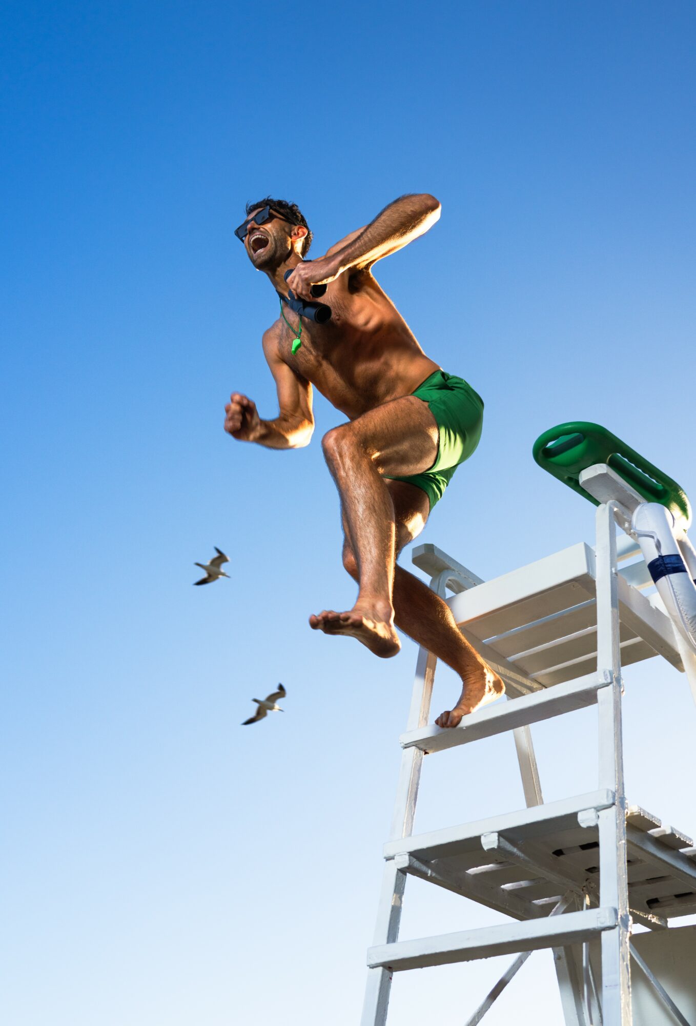 Man jumping off a lifeguard chair into the water, with a clear blue sky and seagulls flying nearby.