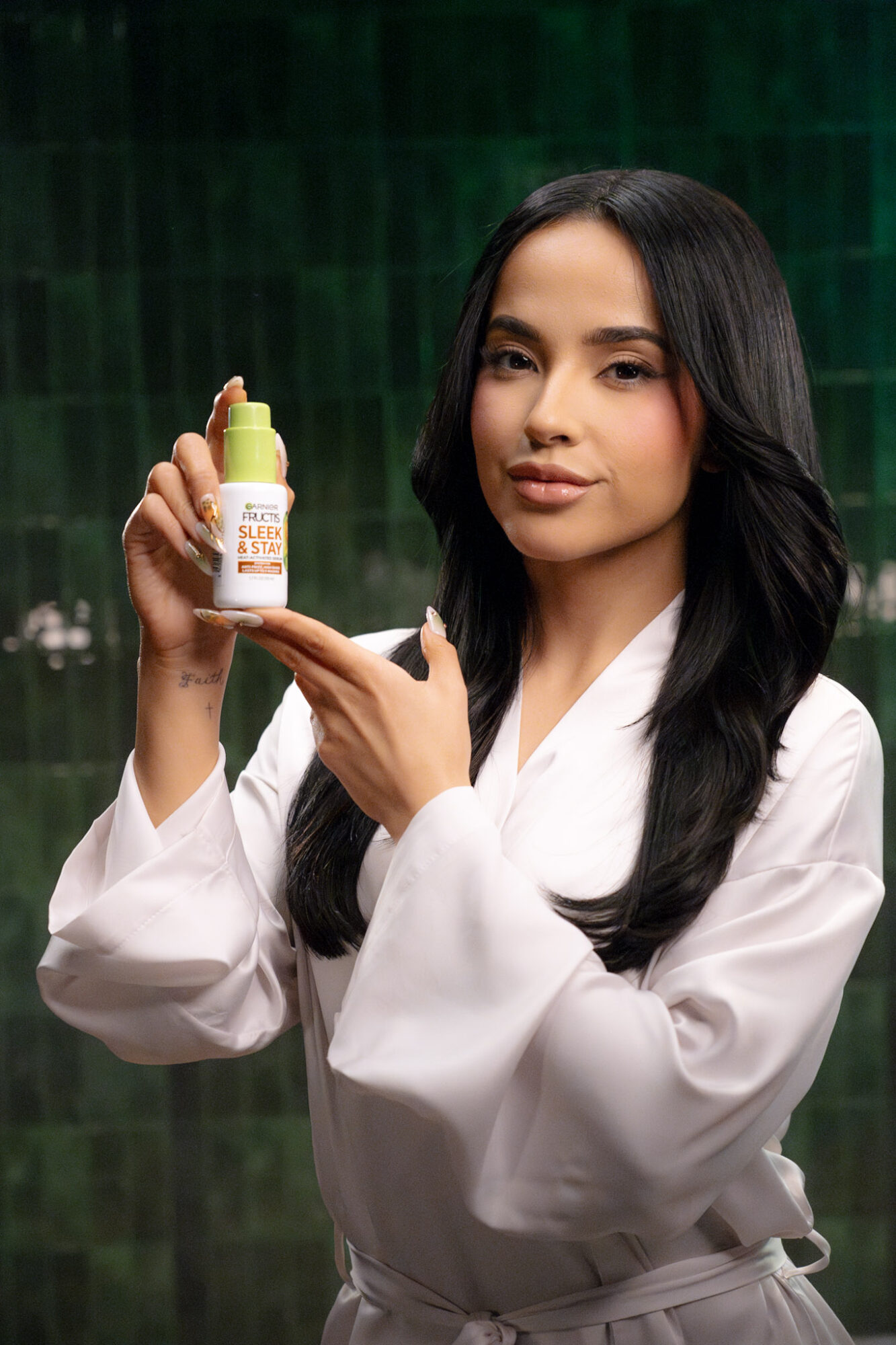 Woman with long black hair in white robe holding a small bottle, pointing at it, against a green background.