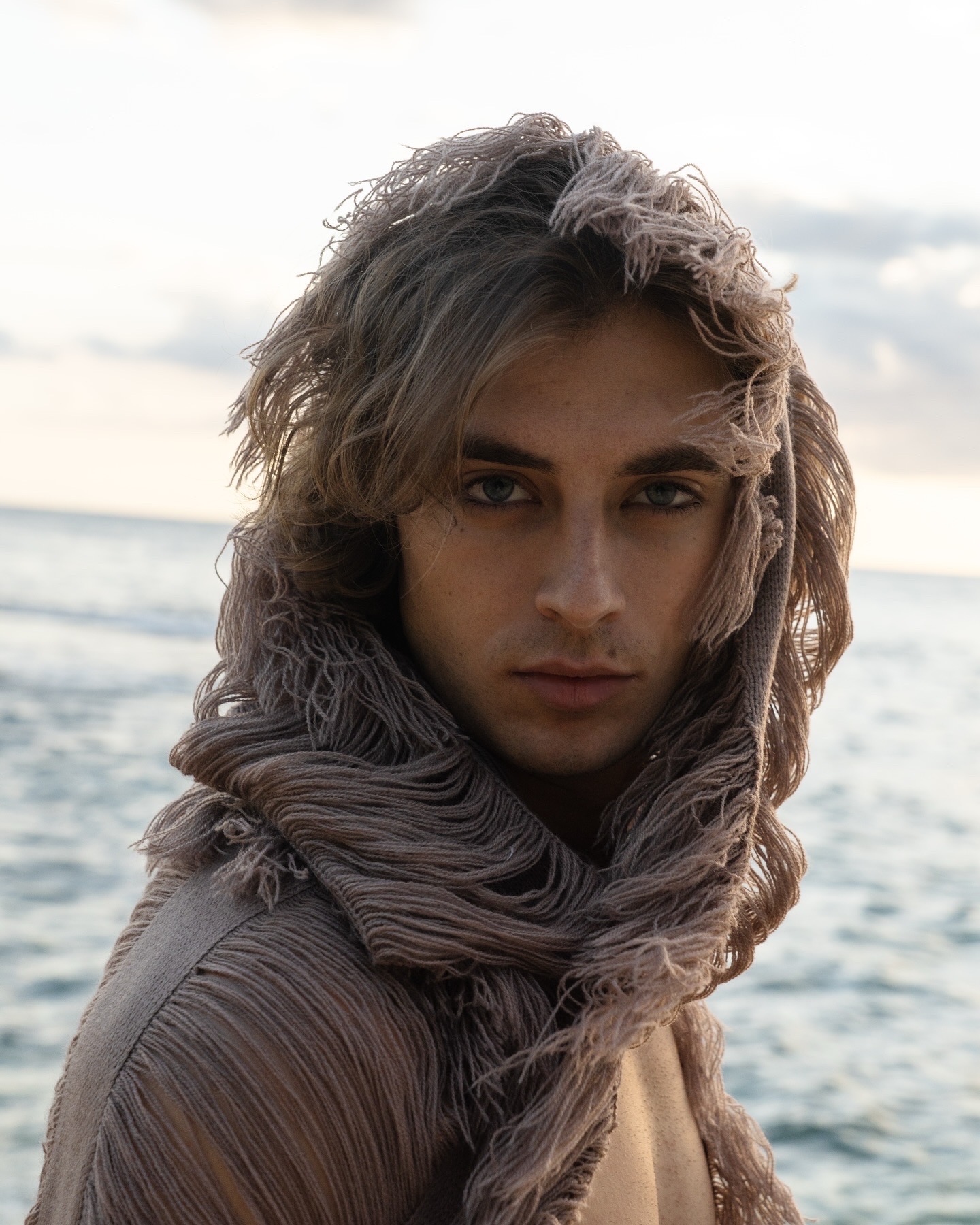 Young person with wavy hair wearing a scarf, standing near the ocean with a cloudy sky background.
