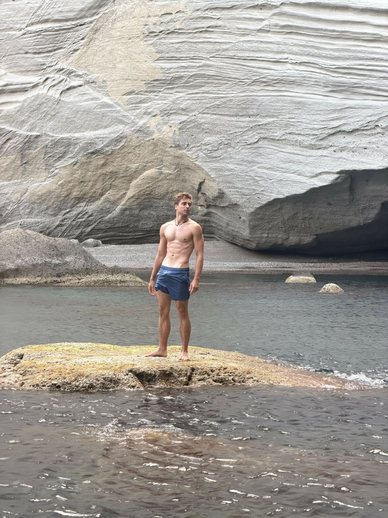 Person standing on a rocky beach near water and cliffs, wearing a swimsuit and shorts, facing sideways.