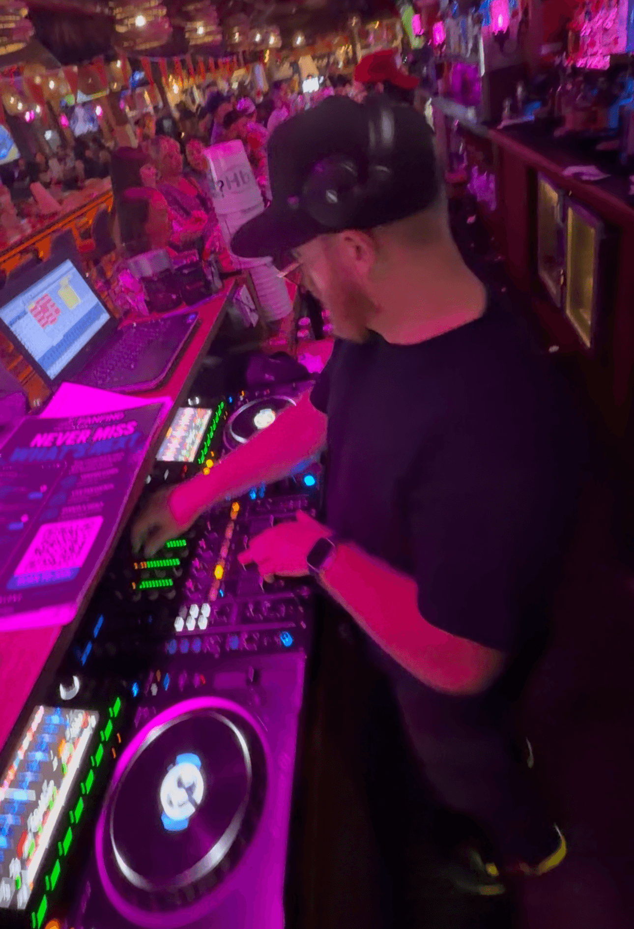 DJ wearing a cap and black shirt operates a mixing console in a colorful, lively nightclub with a crowd in the background.