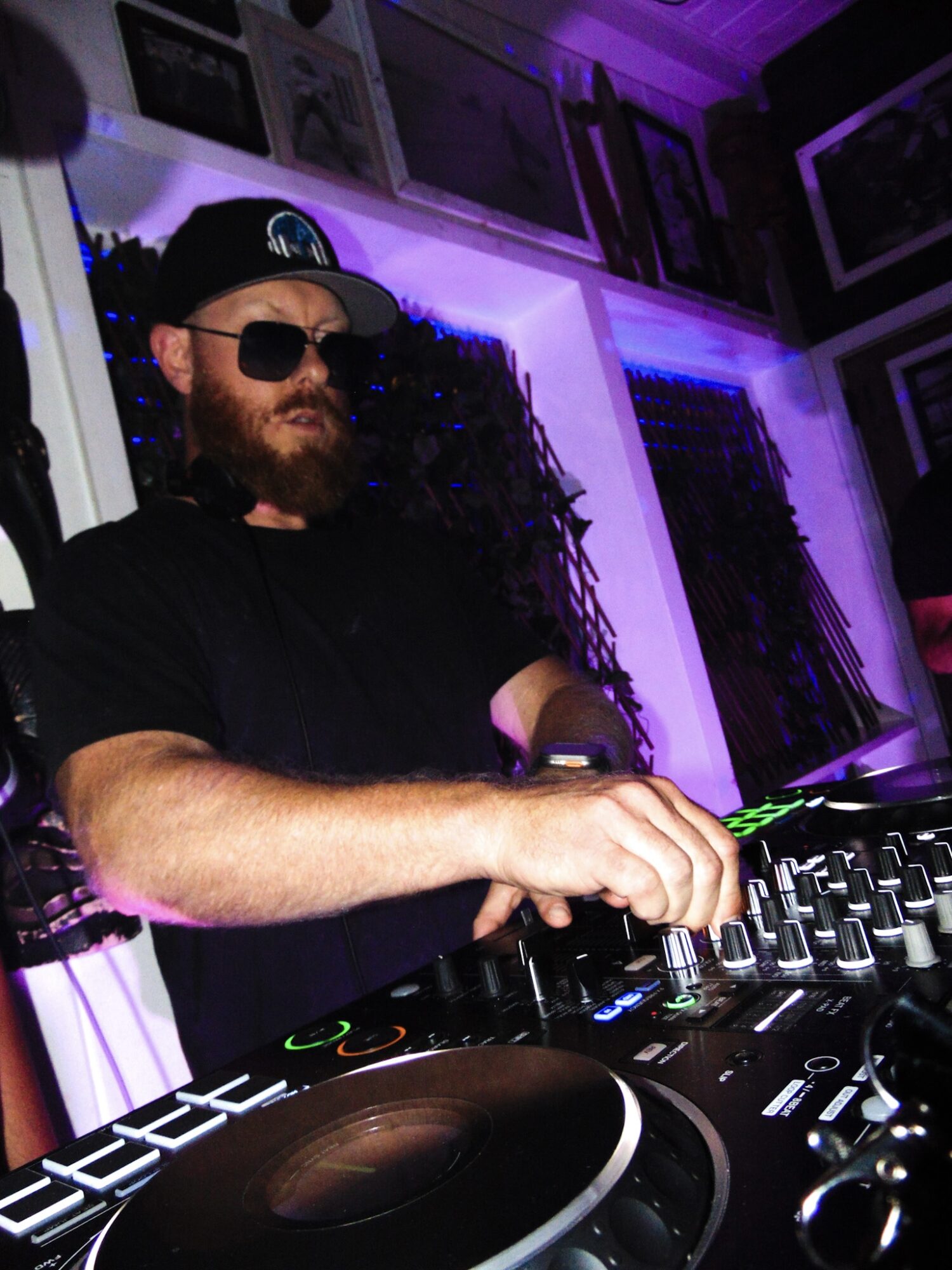 Bearded man wearing sunglasses and a cap DJing with equipment in a dimly lit room with purple lighting.