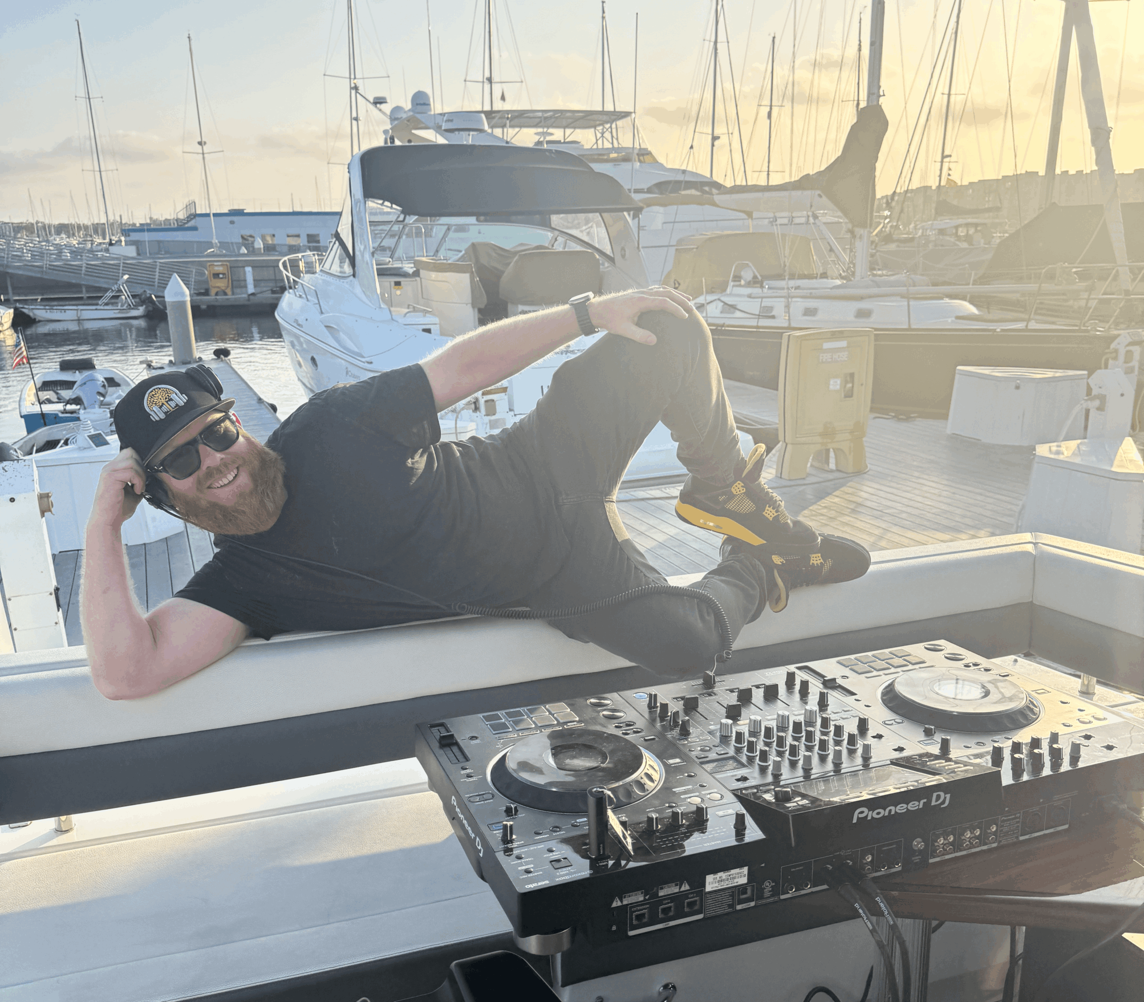 Person lying on a boat deck with a DJ controller, marina with boats in background, sunset sky, person wearing sunglasses and black clothing.