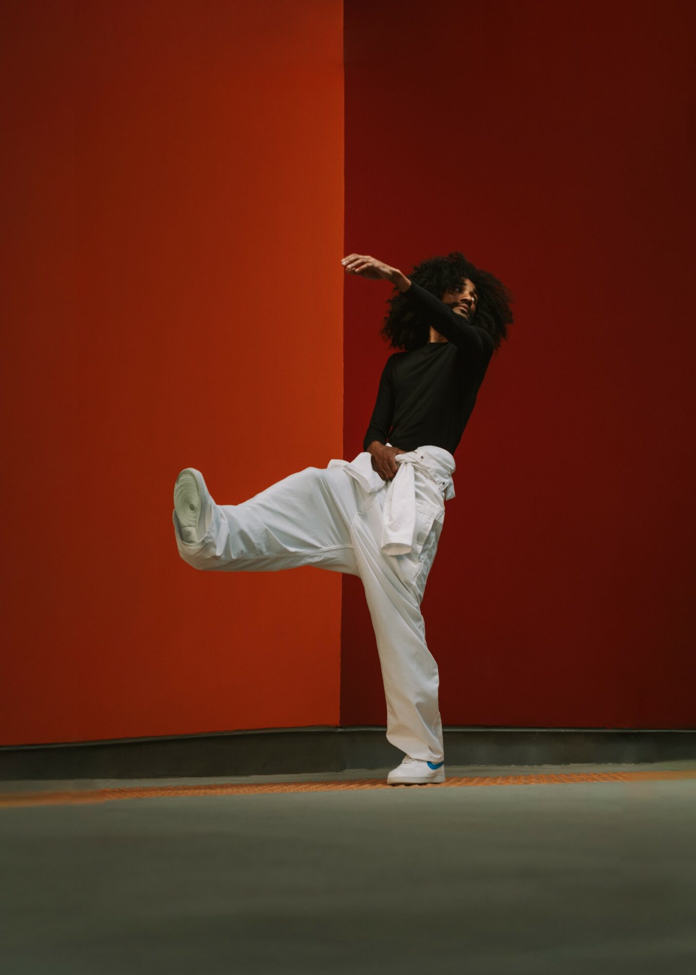 Person with curly hair in black top and white pants performs a dance move against a red wall.