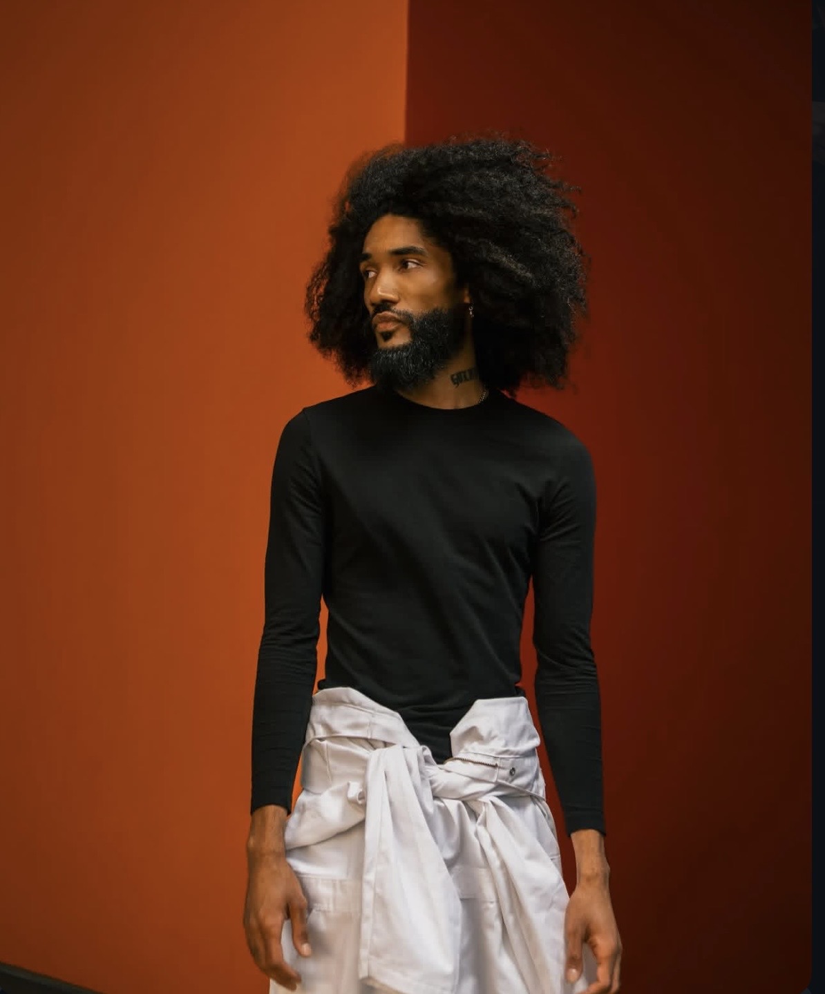 Person with curly hair wearing a black long-sleeve shirt and white pants tied at the waist, standing against an orange background.