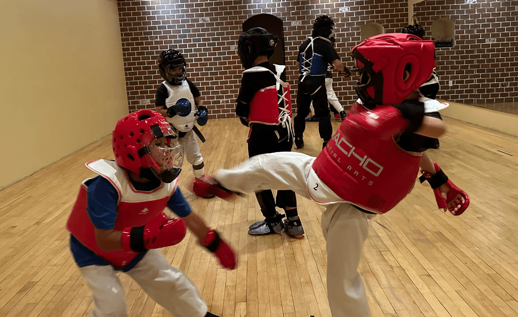 Two children practicing martial arts with protective gear, one executing a kick, others watching in a gym.