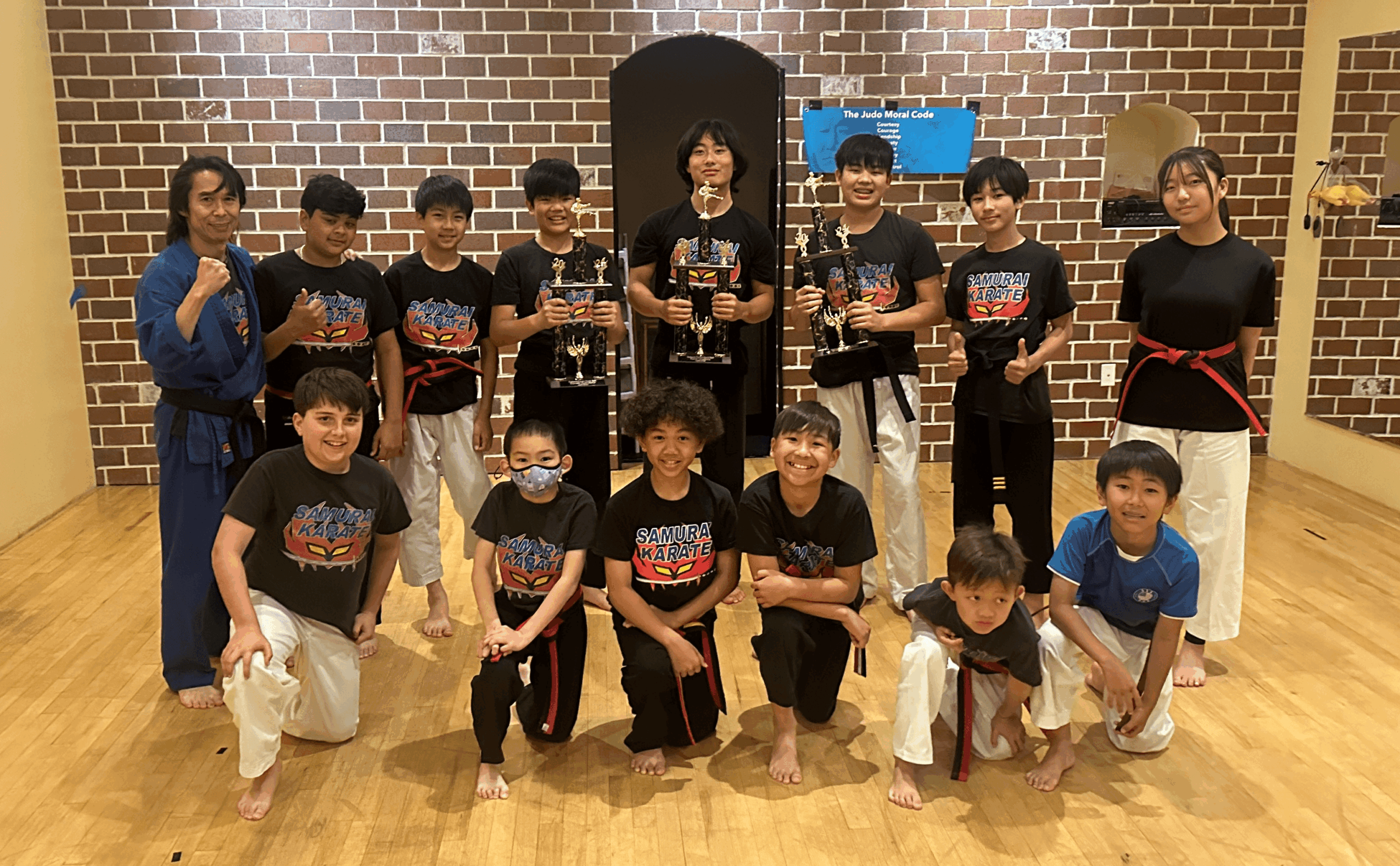 Group of children and two adults in martial arts uniforms and casual clothes, standing and kneeling indoors against a brick wall.