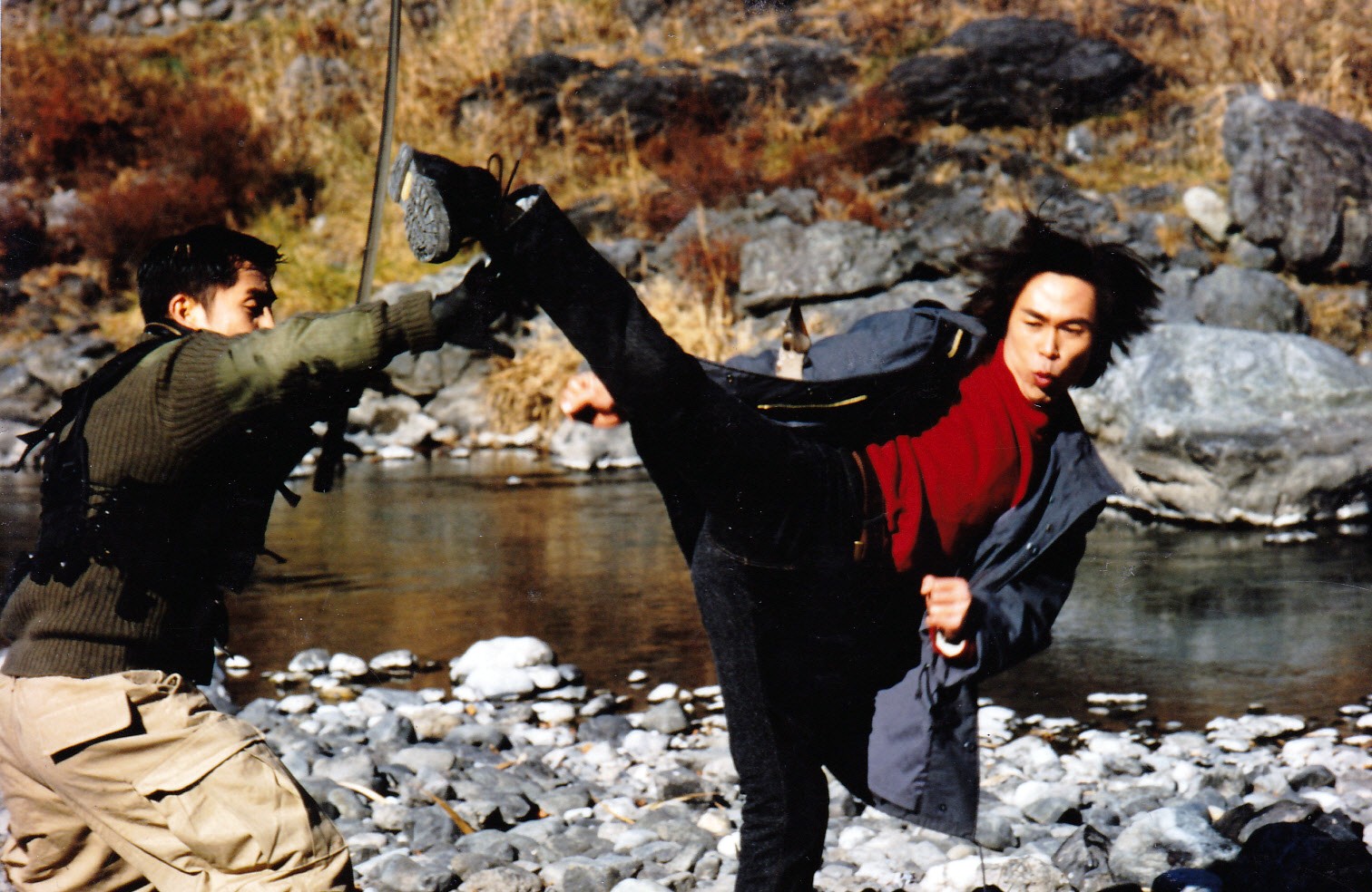 Two people fighting outdoors near a river, one person kicking the other, rocky landscape background.