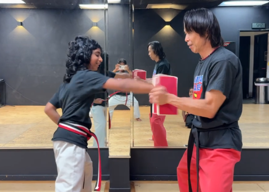 Two people practicing martial arts in a studio, one holding pads and the other performing a punch, with a mirror behind them.