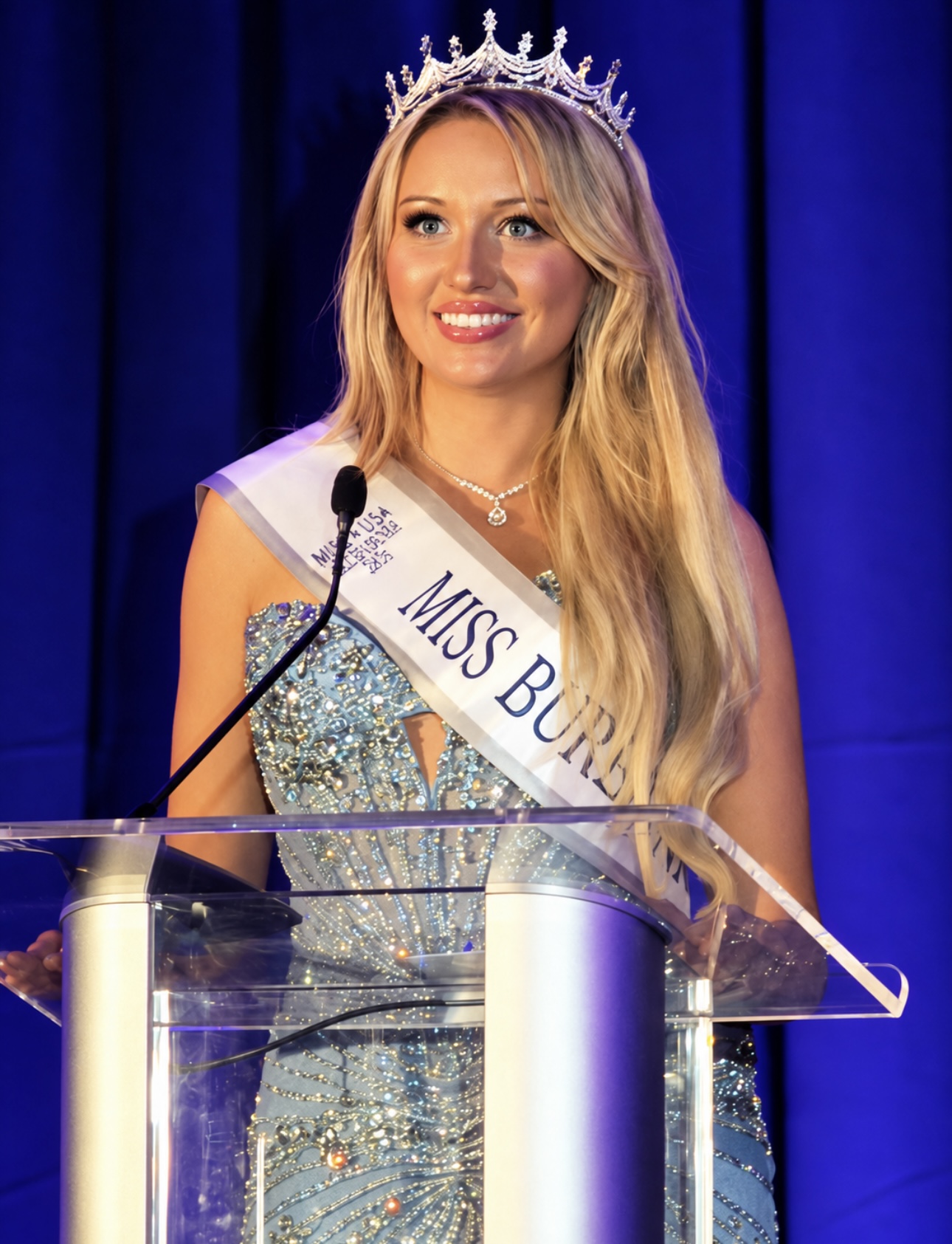 Young woman wearing a crown and sash, smiling at a podium during a pageant event, with a blue curtain background.