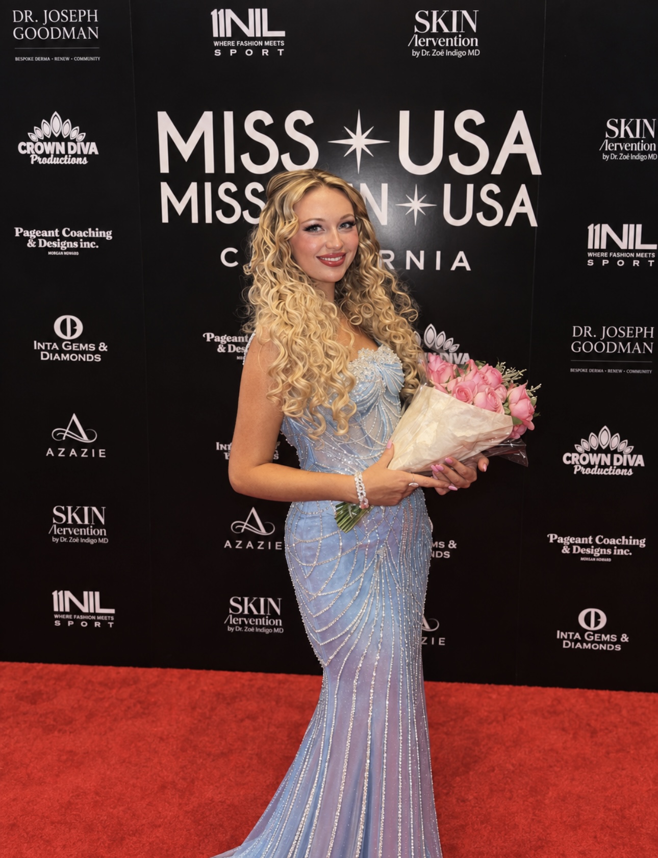 Woman with long curly blonde hair in a blue gown holding a bouquet of pink flowers on red carpet, black backdrop with event logos