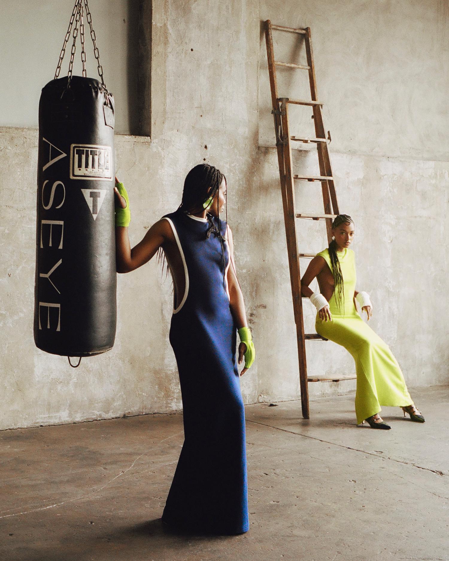 Two women in a room with a punching bag and a ladder; one standing and the other sitting on the floor.