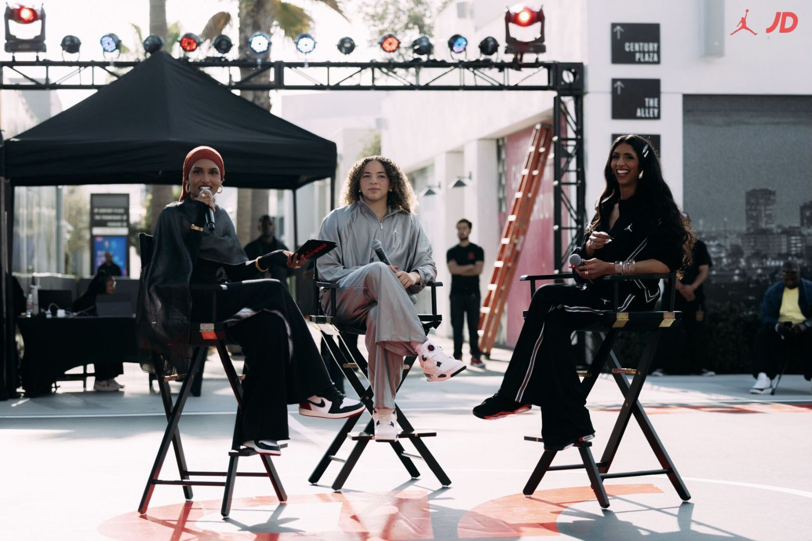 Three women sit on chairs outdoors under a black canopy, with a cityscape and people in the background.