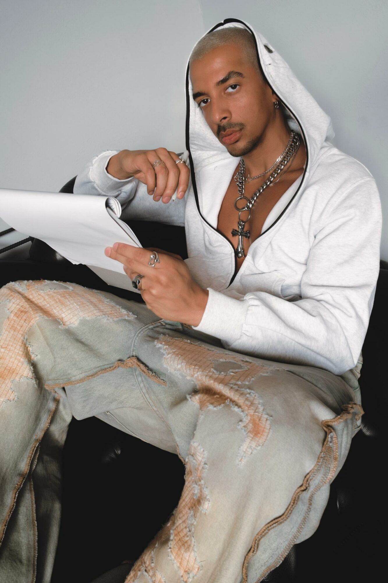 Man with a shaved head and beard wearing a white hoodie, holding papers, sitting on a distressed jeans, against a plain background.