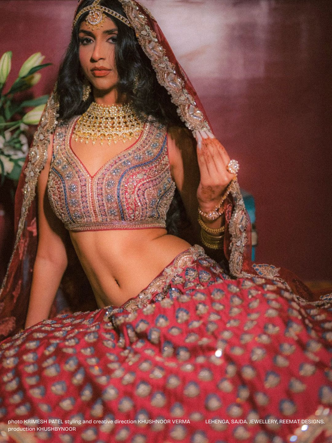 Woman in traditional attire with jewelry, sitting against a plain background, looking at the camera.