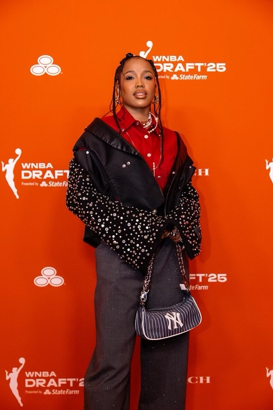 Woman with braided hair wearing a black and red jacket, holding a purse, standing against an orange background with event logos.
