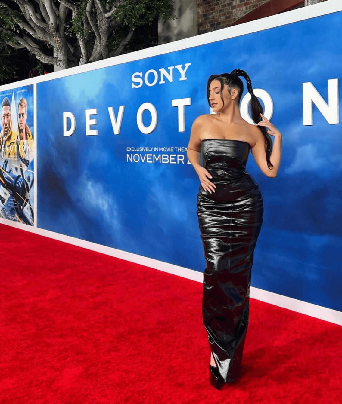 Woman in a strapless black gown standing on red carpet in front of a blue promotional wall for Sony's Devotion movie.