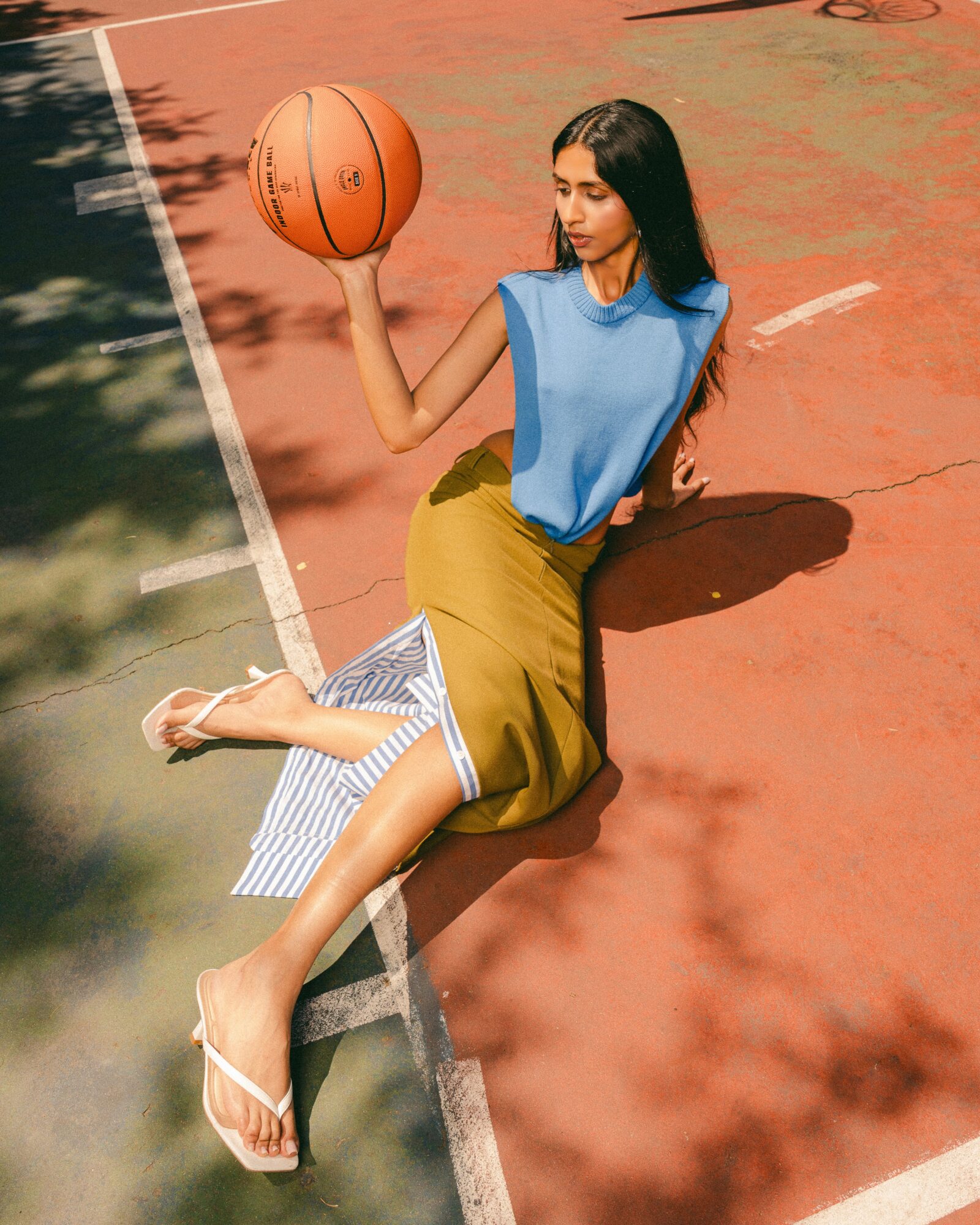 Woman sitting on a basketball court holding a basketball, wearing a blue top and yellow skirt, with shadows on the court.