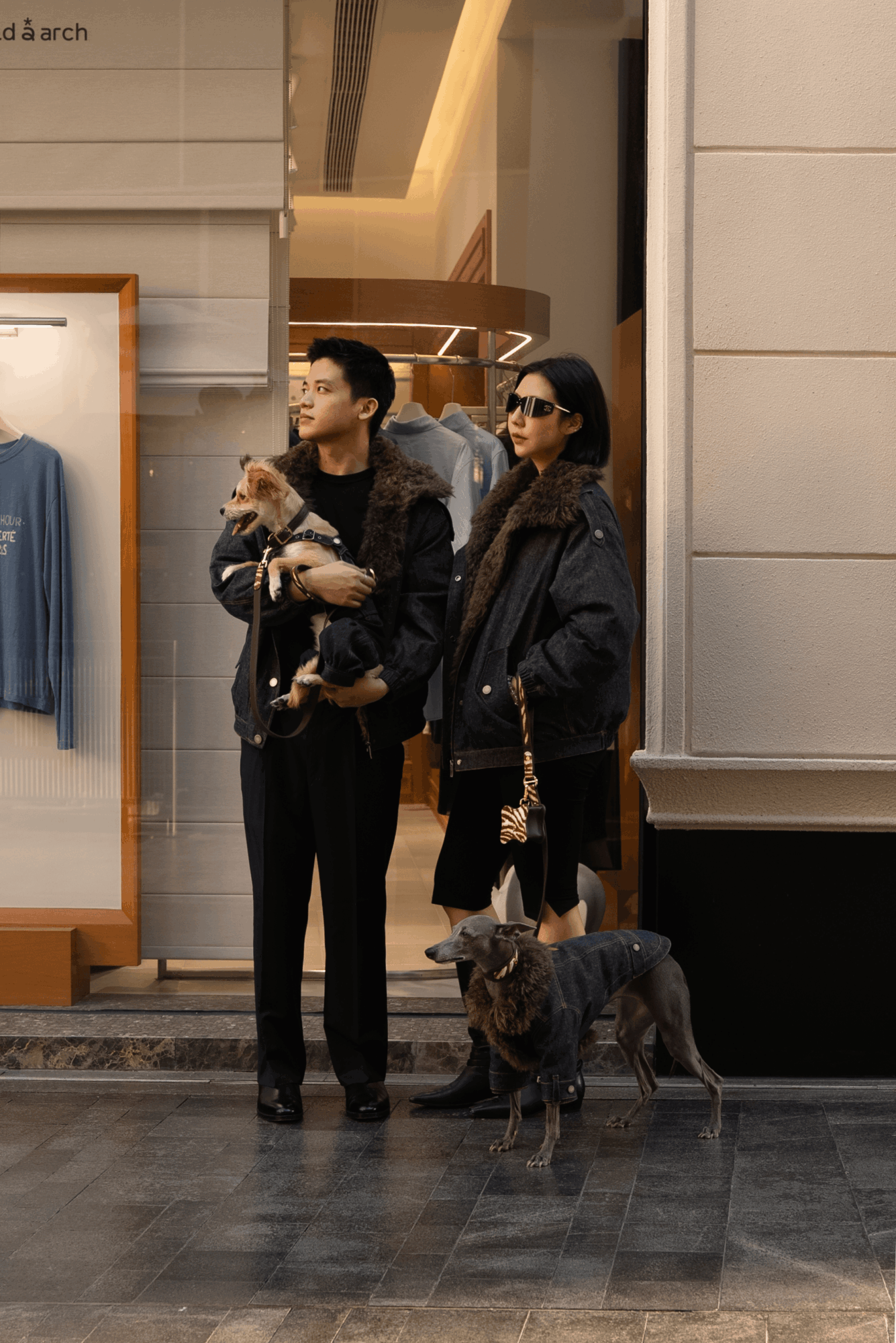 Two women stand outside a building, one holding a small dog, with a larger dog on a leash nearby, in an urban setting.