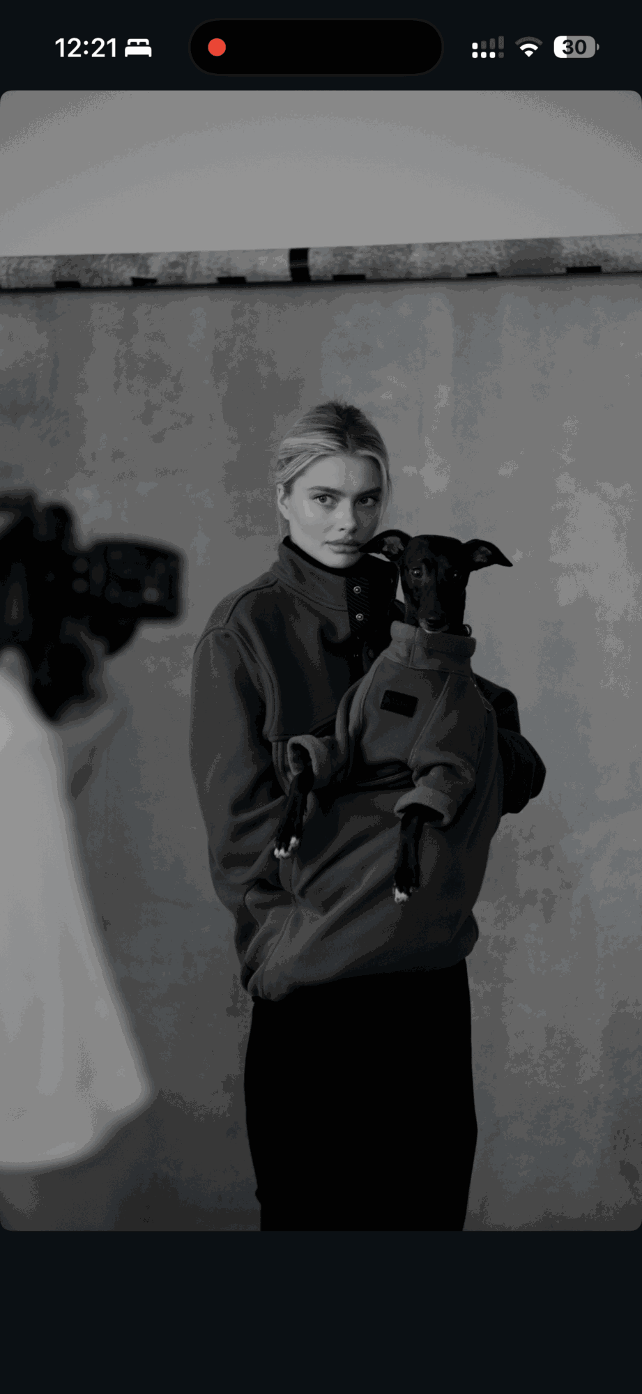 Person holding a dog, standing against a wall, in black and white, with a camera in the foreground.