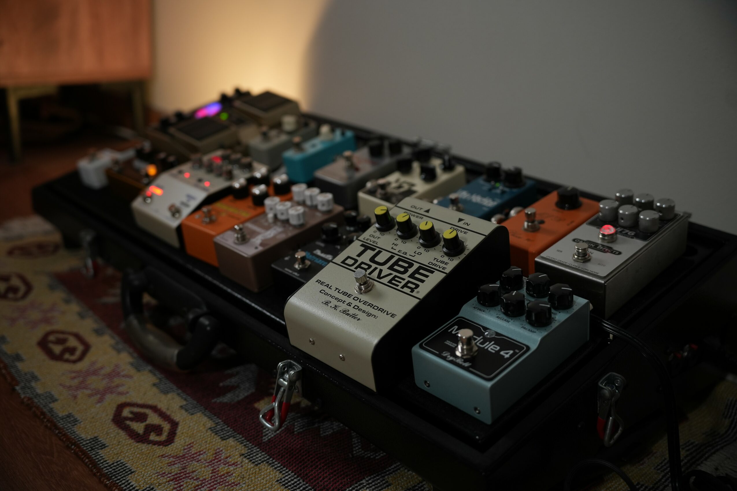 Multiple guitar effect pedals arranged on a black pedalboard, placed on a patterned rug in a room.