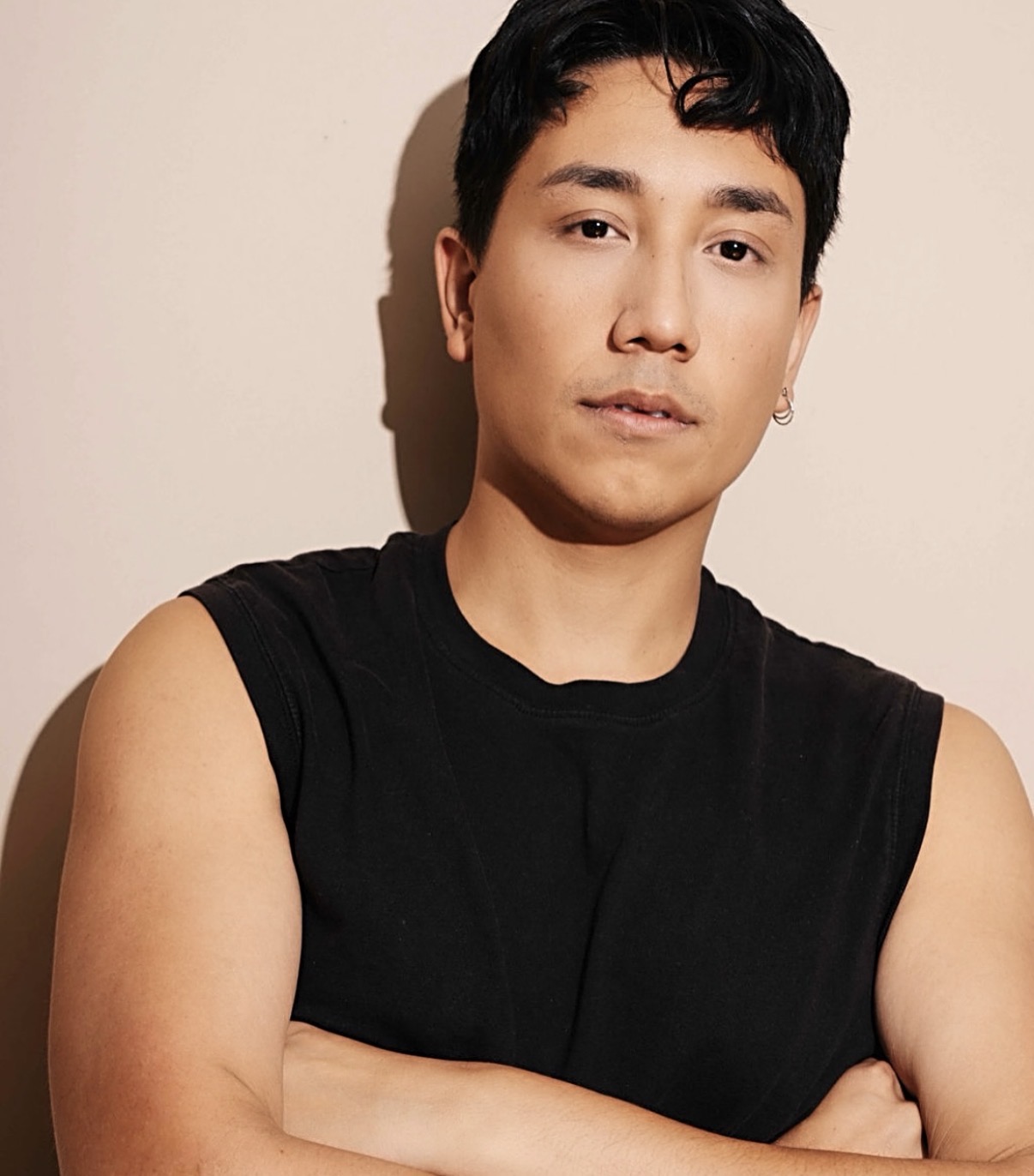 Young person with short black hair, wearing a sleeveless black shirt, standing against a plain beige background.