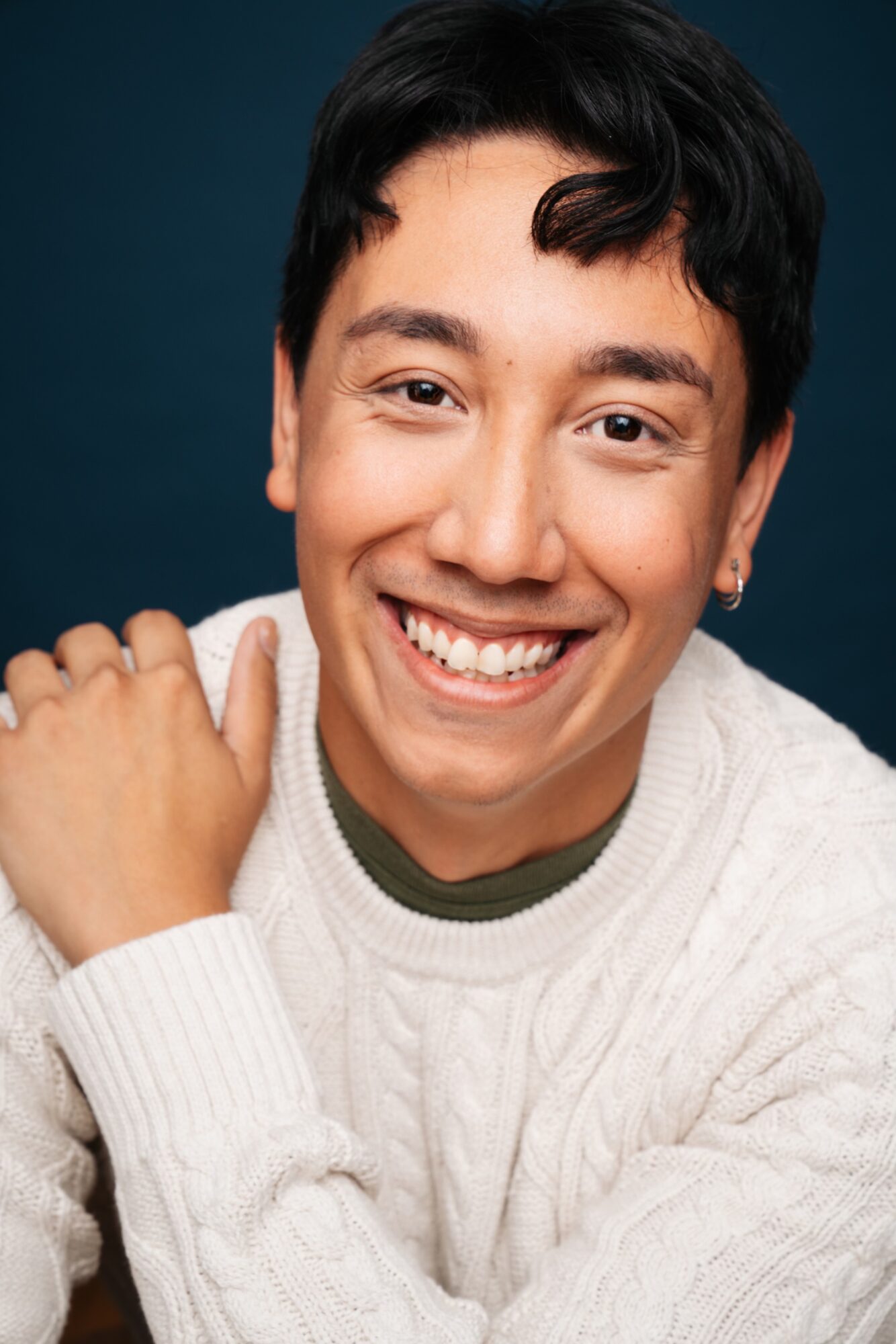 Young man with dark hair smiling, wearing a white sweater, against a dark background.