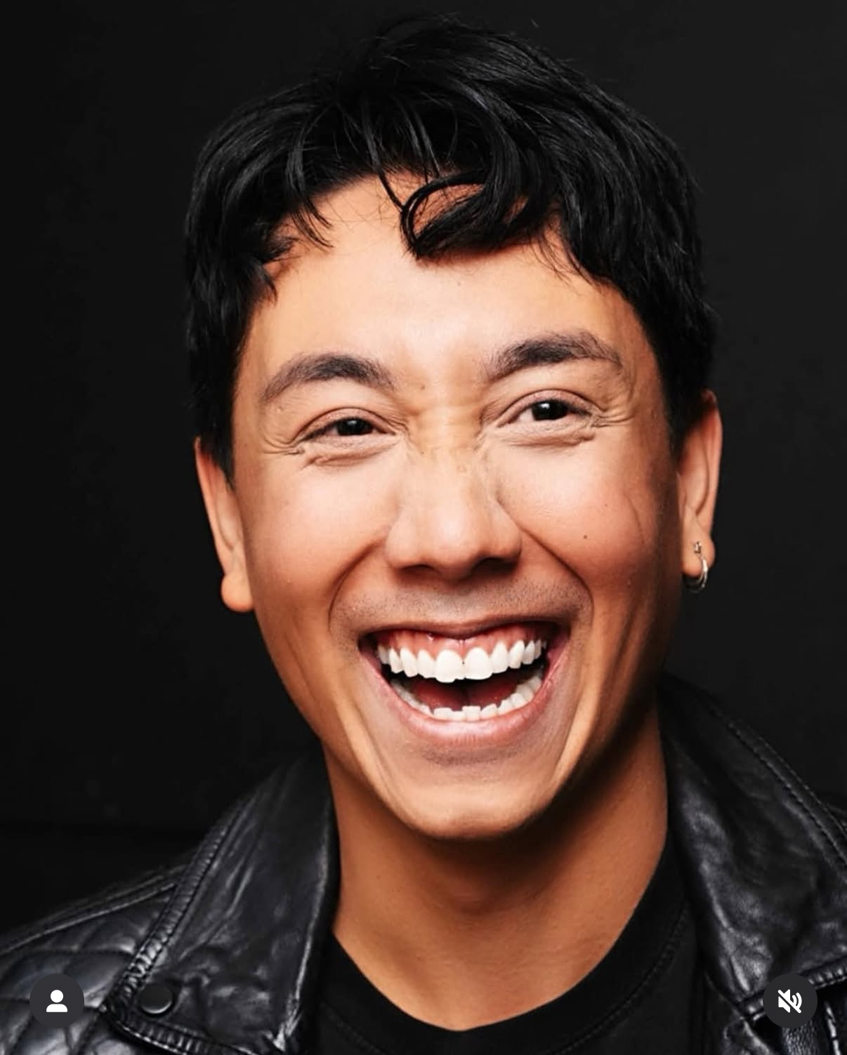 Person with short black hair smiling broadly, showing teeth, wearing a black leather jacket, against a dark background.