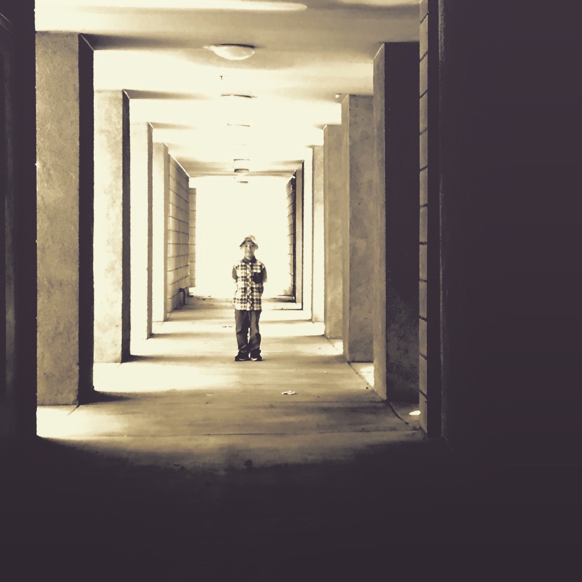 Person standing in a corridor with repeating archways, bright light at the end, shadows on the floor, black and white image.