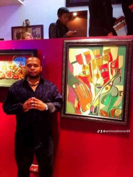 Person standing in front of colorful abstract painting, hands clasped, in an indoor setting with other people and artwork visible.