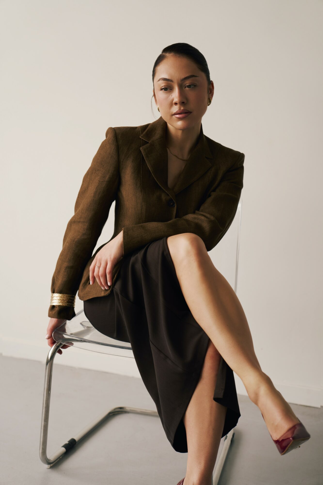 Woman sitting on a chair with crossed legs, wearing a brown blazer and black skirt, against a plain background.