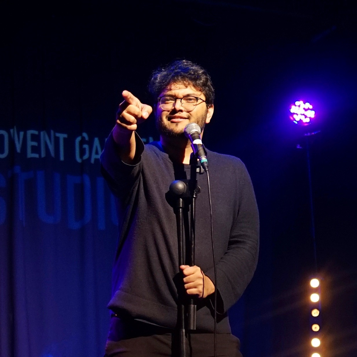 Man with glasses and beard points forward on stage with microphone, purple stage light in background.