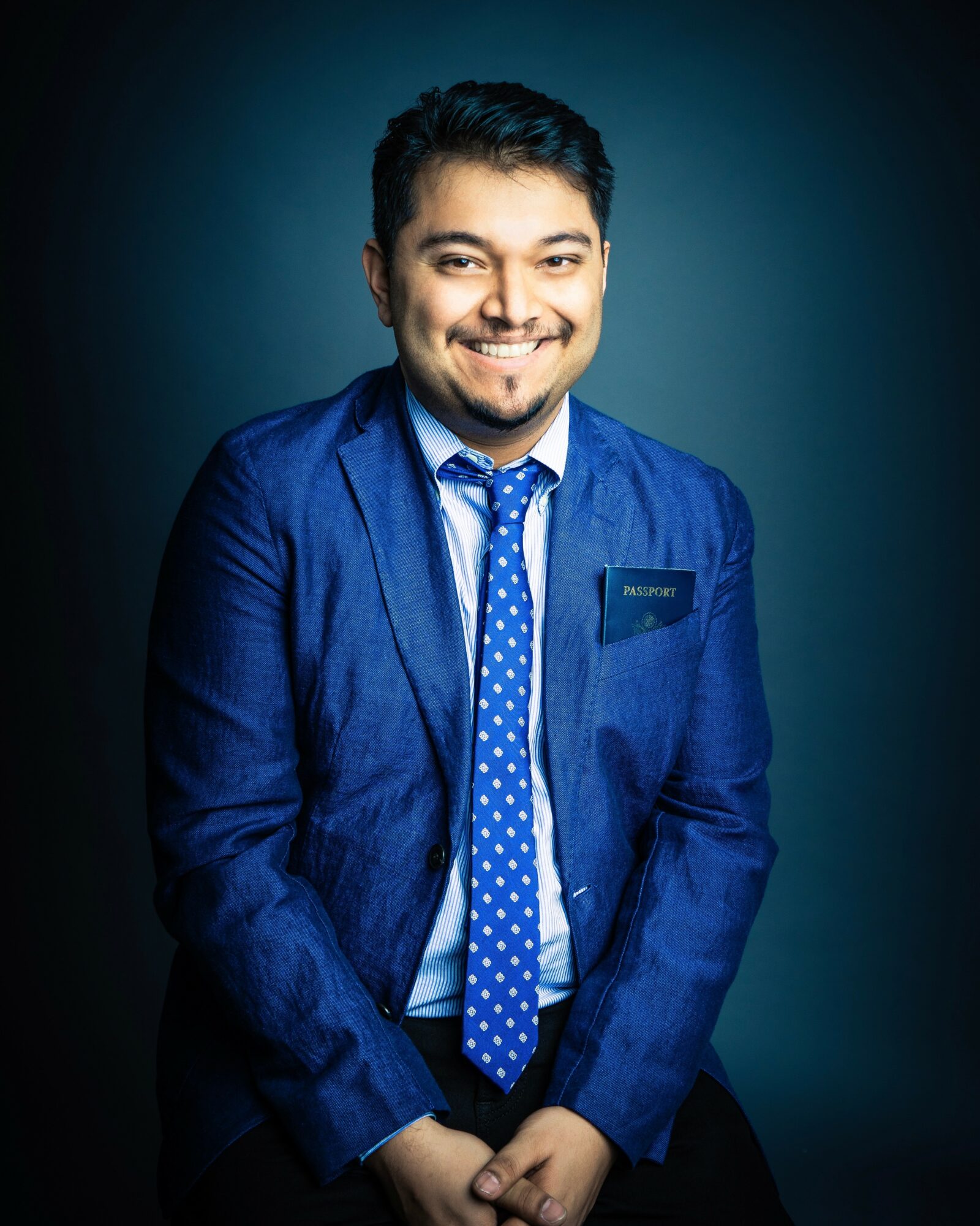 Smiling man in a blue suit and polka dot tie, standing against a dark background.