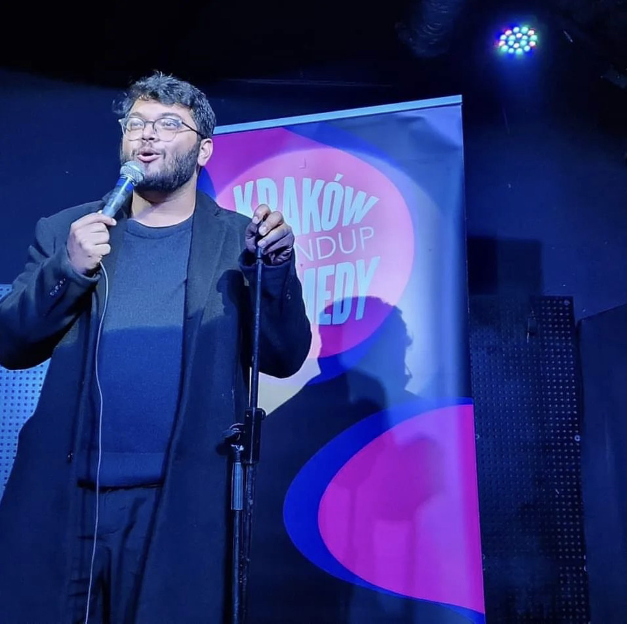 Man with glasses and beard speaking into a microphone on stage, with a colorful backdrop and stage lighting.