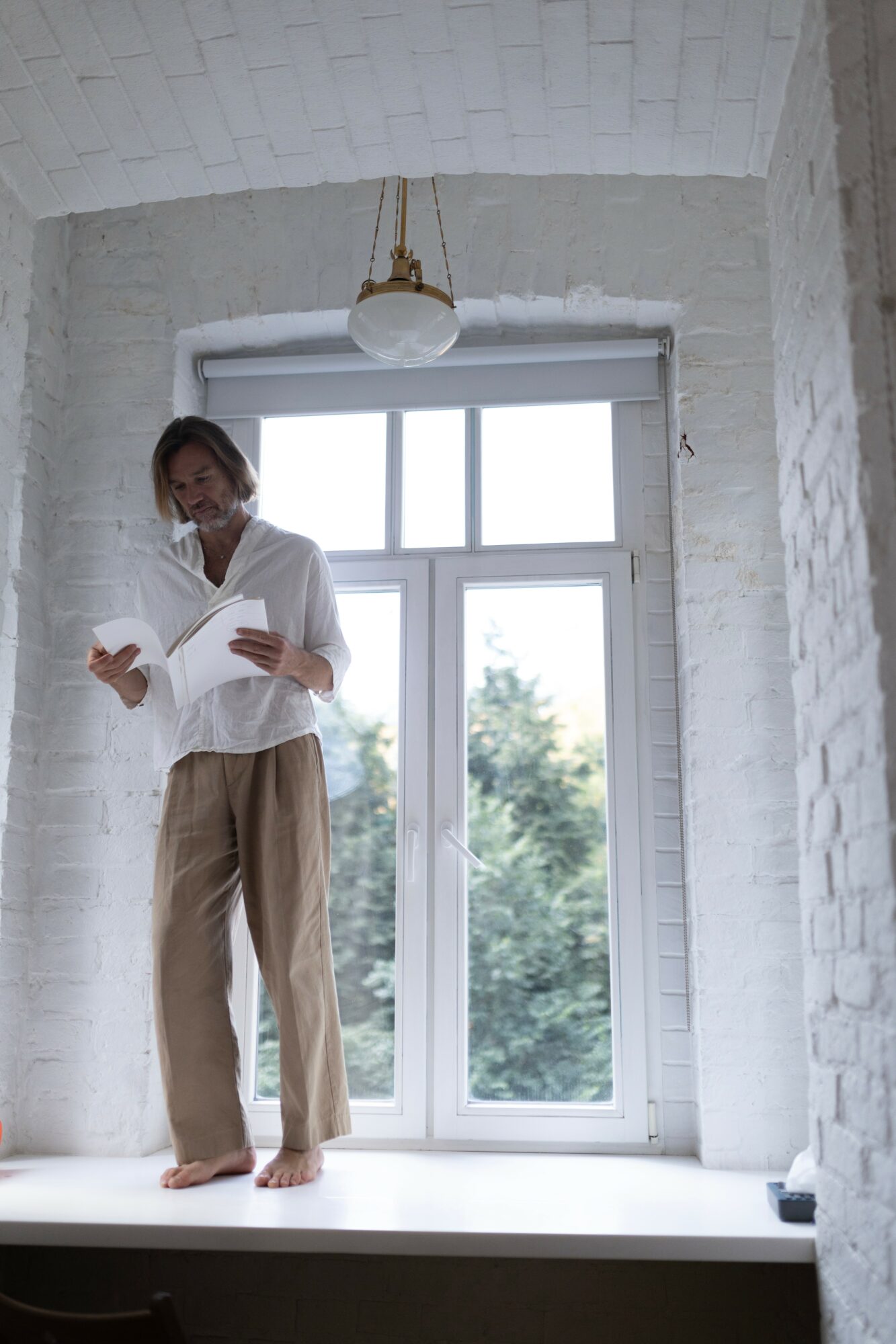 Person standing on windowsill reading a book, window showing trees outside, white brick walls, hanging light fixture, barefoot.