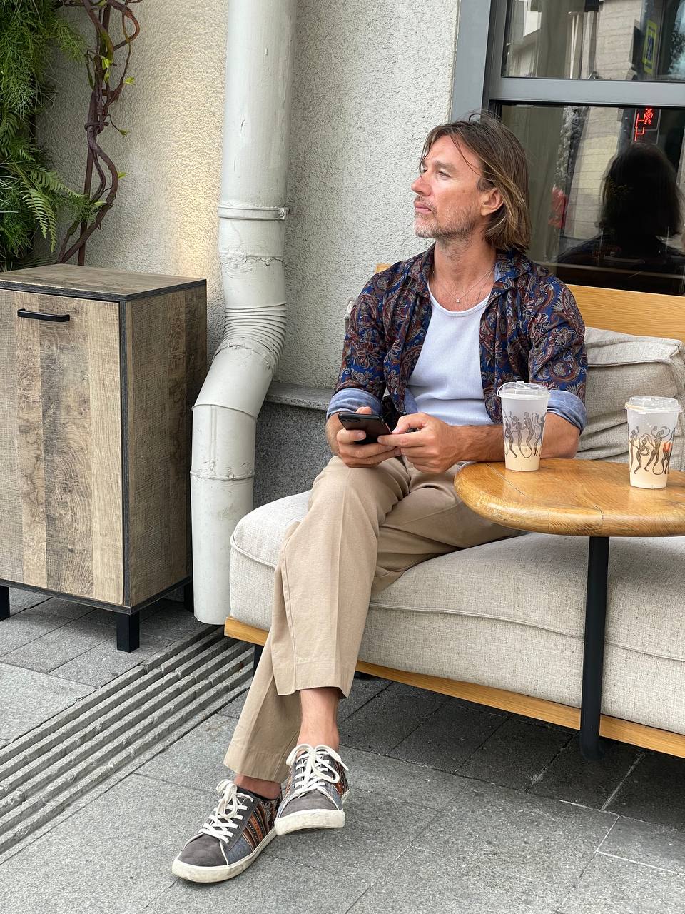 Man sitting on a sofa outside, holding a phone, with a table and drinks nearby.