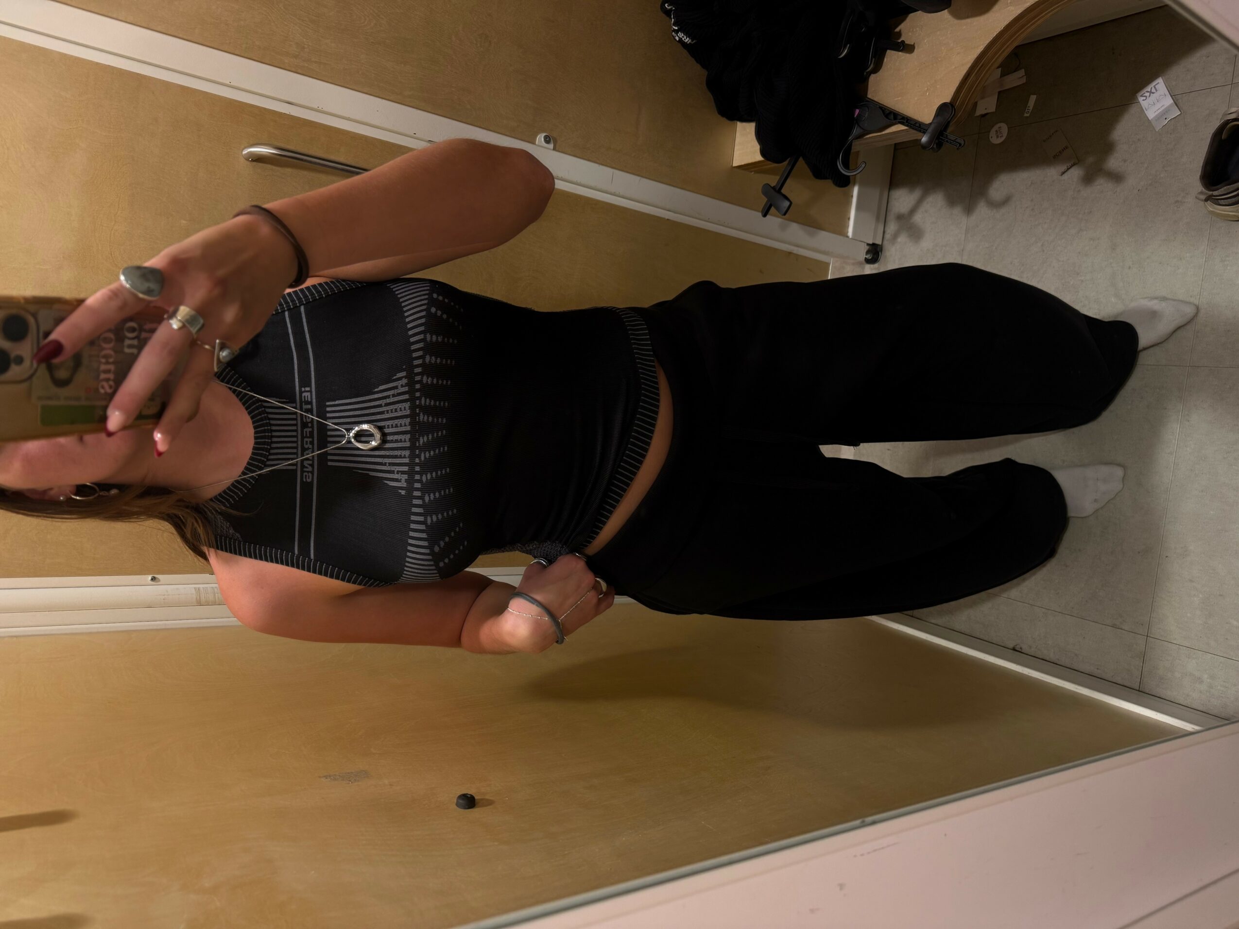 Person taking mirror selfie in a room, wearing black pants and a black top, holding phone with left hand, face not visible.