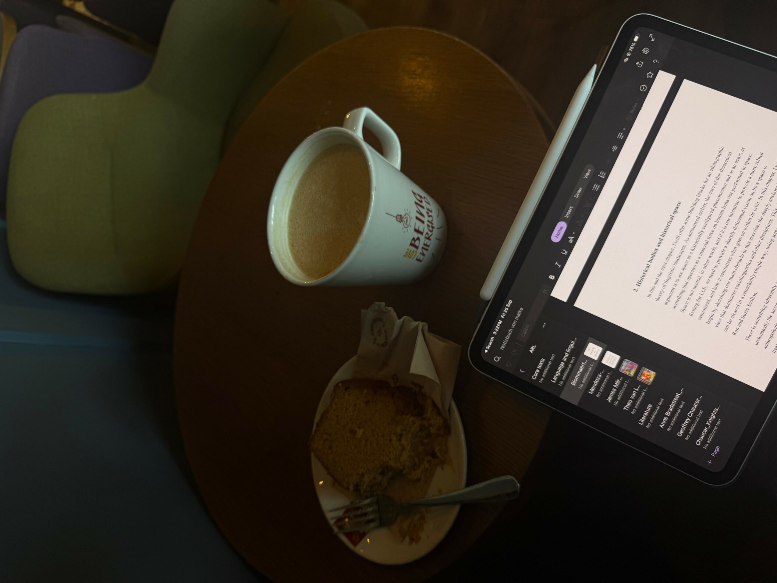 Coffee mug, plate with cookie, and tablet on a wooden table, with a green chair nearby.