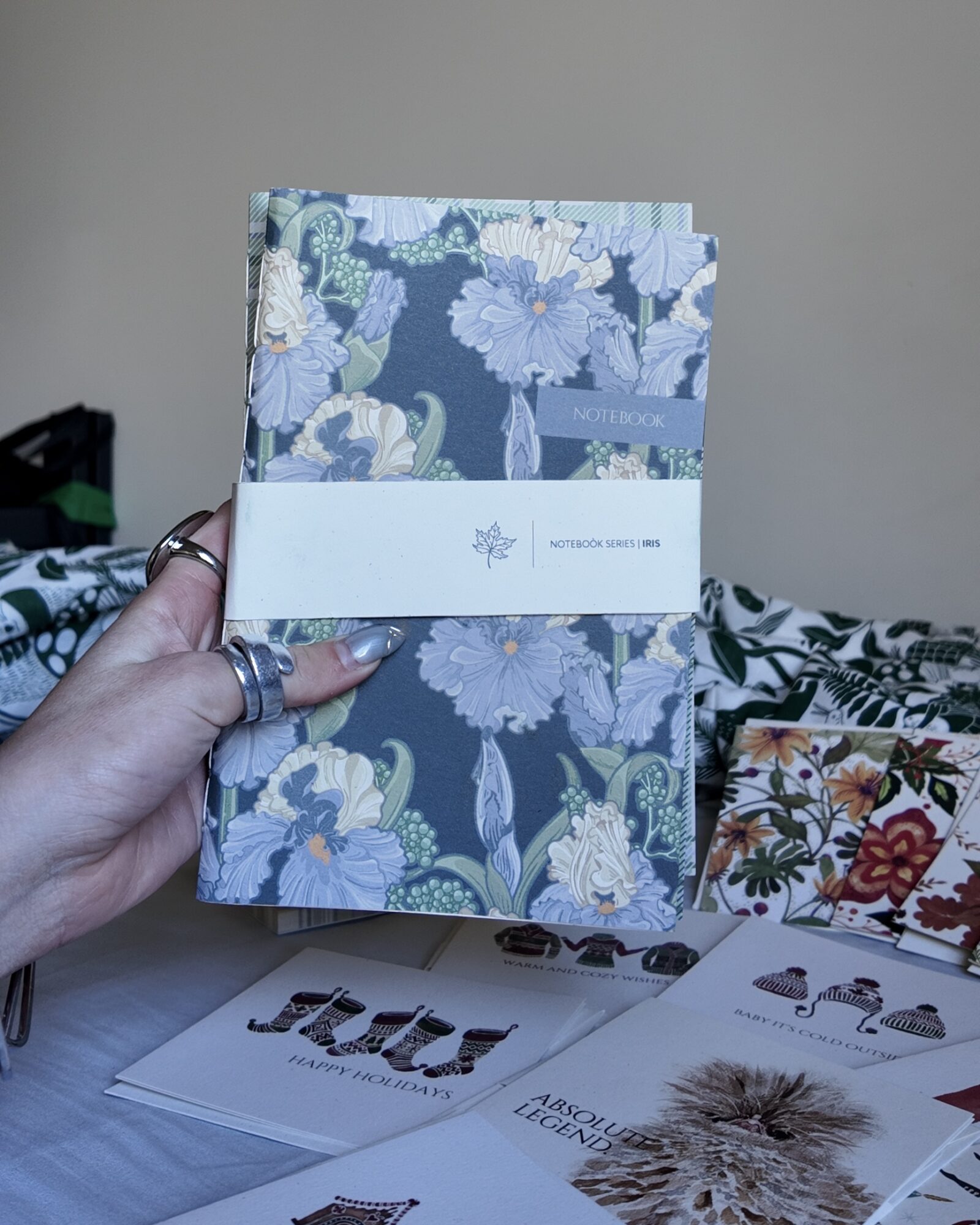 Hand holding a floral-patterned book with a white band, background includes patterned fabric and papers on a table.