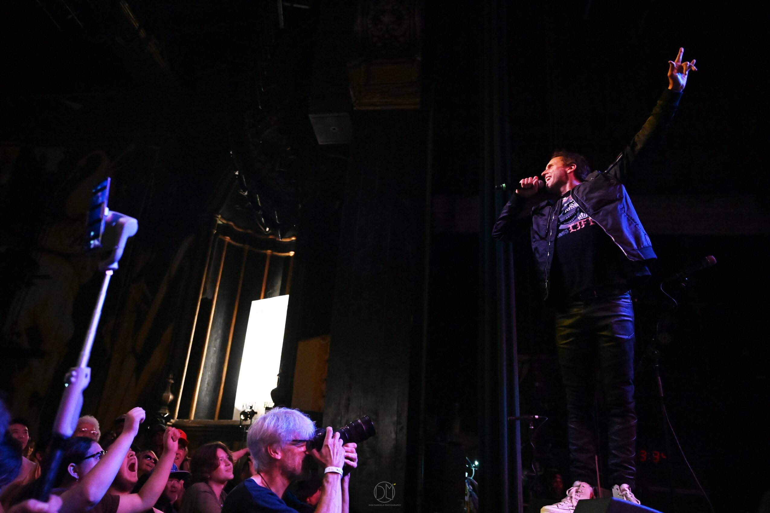 Performer on stage singing with one arm raised, audience clapping and cheering, dark background, stage lighting, and a large screen.